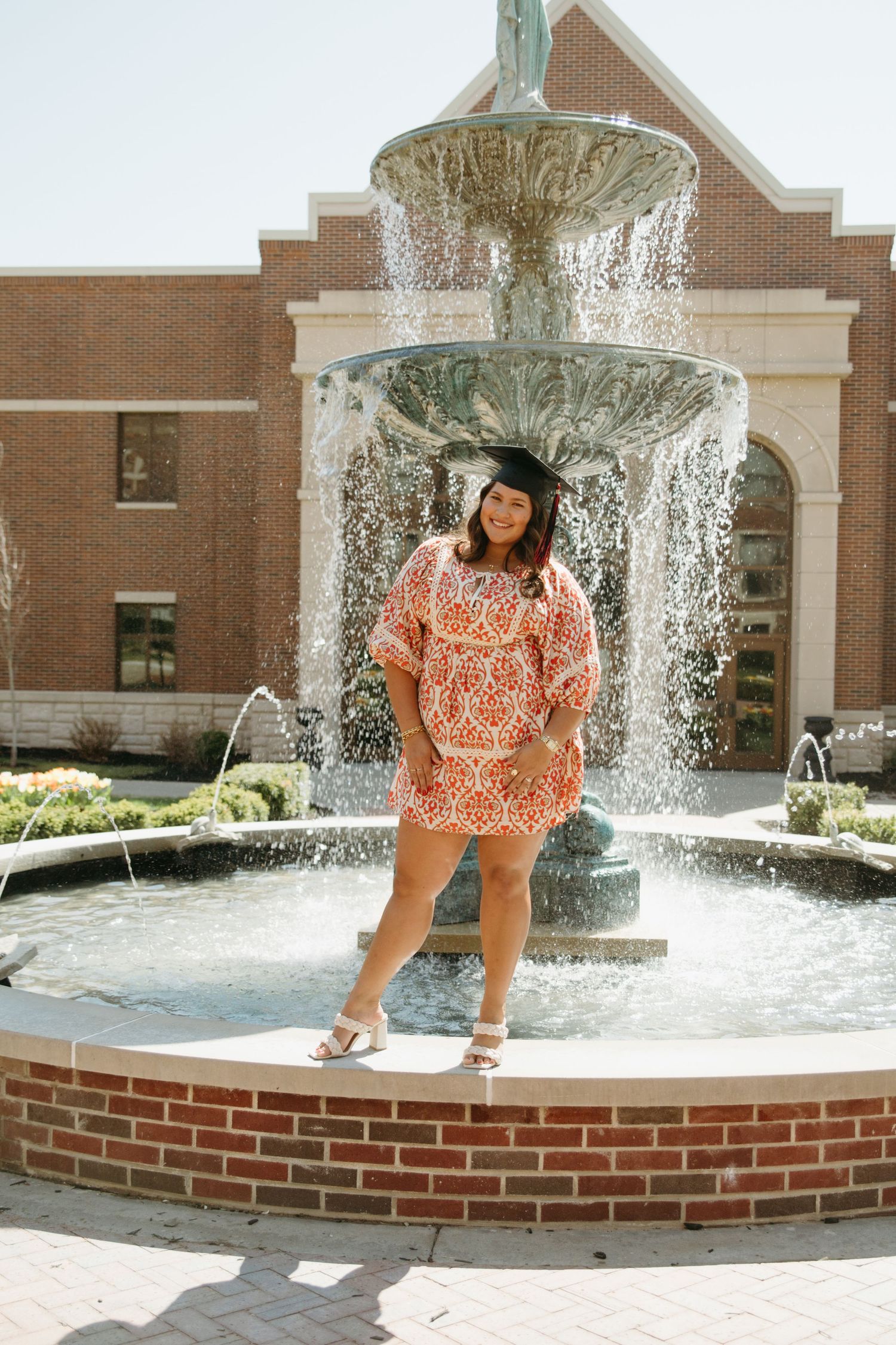 Graduation portrait of a smiling woman in cap and red patterned dress by a campus fountain.
