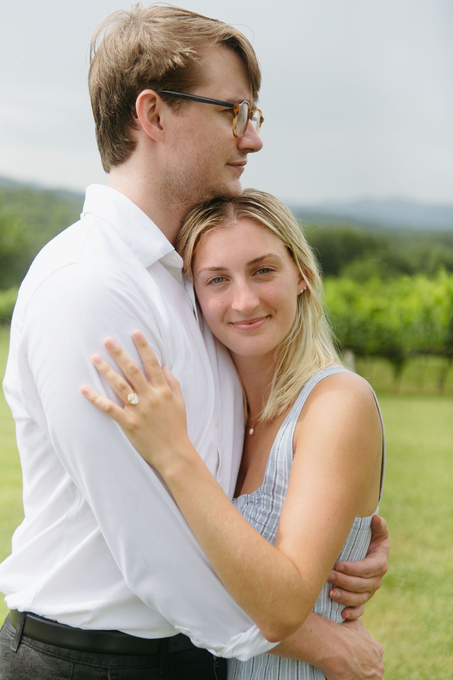 Engaged couple hugging closely outdoors at vineyard, woman showing diamond engagement ring.