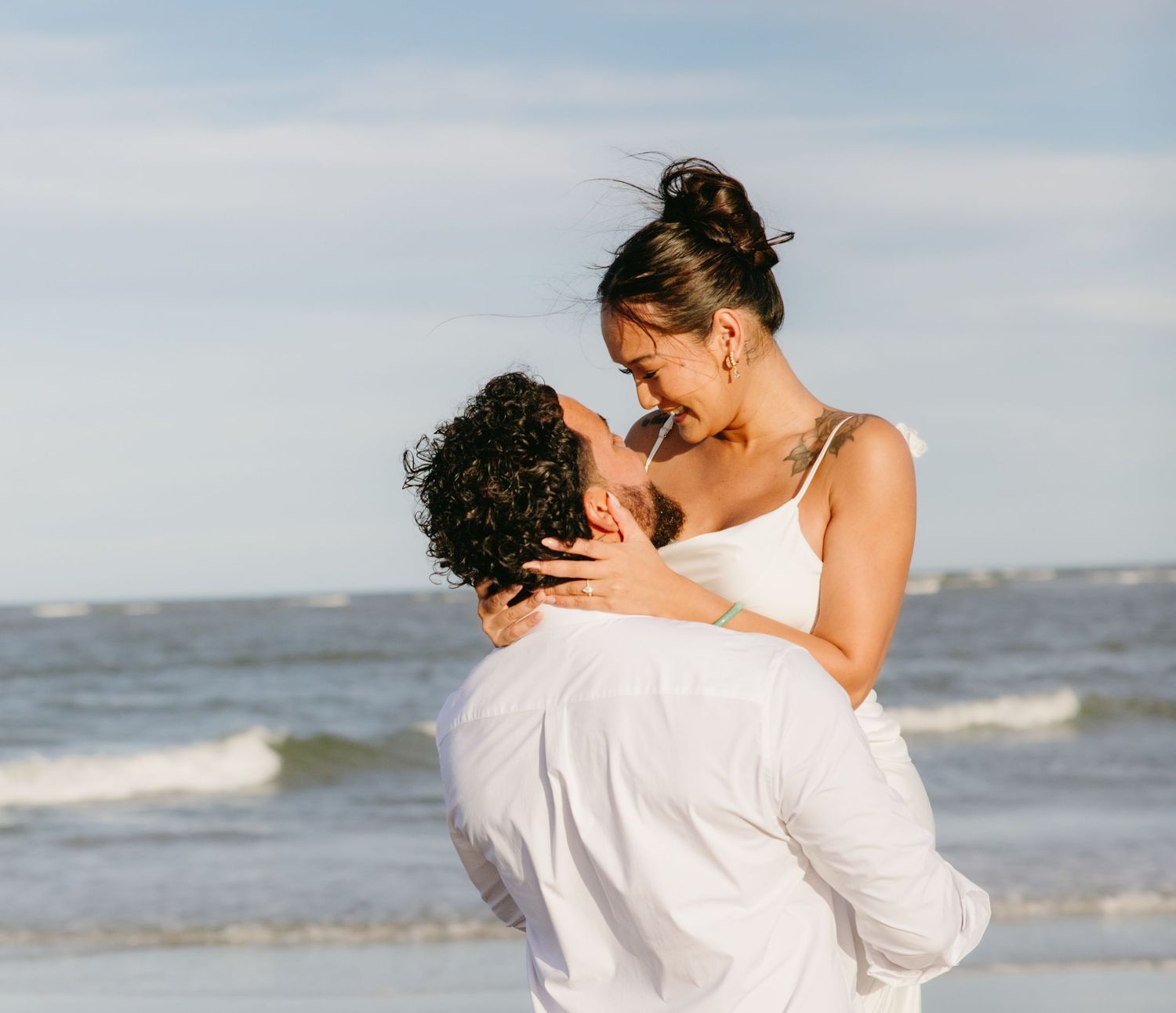 Man lifting woman in white dress on beach as they smile at each other.
