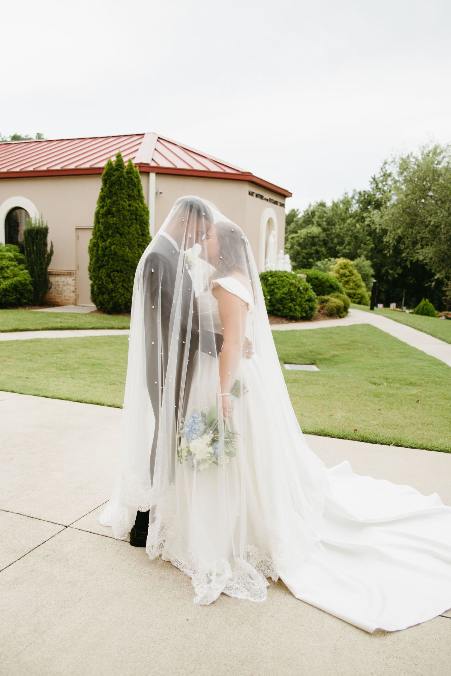 Bride and groom share intimate kiss hidden beneath flowing pearl-adorned wedding veil outside elegant venue.
