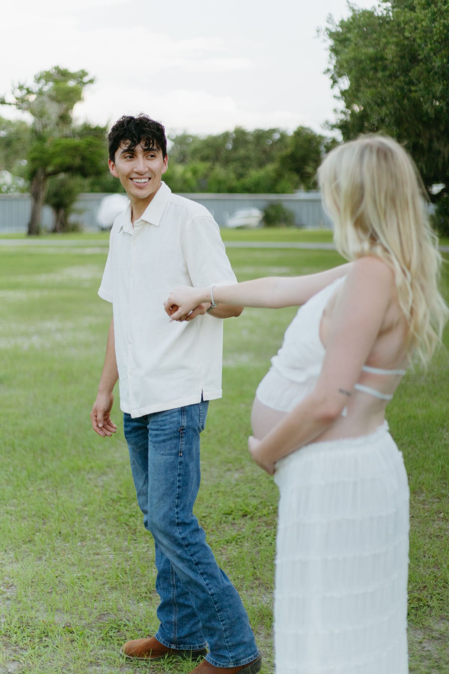 Smiling man in white shirt holding hands with pregnant woman in white dress outdoors.