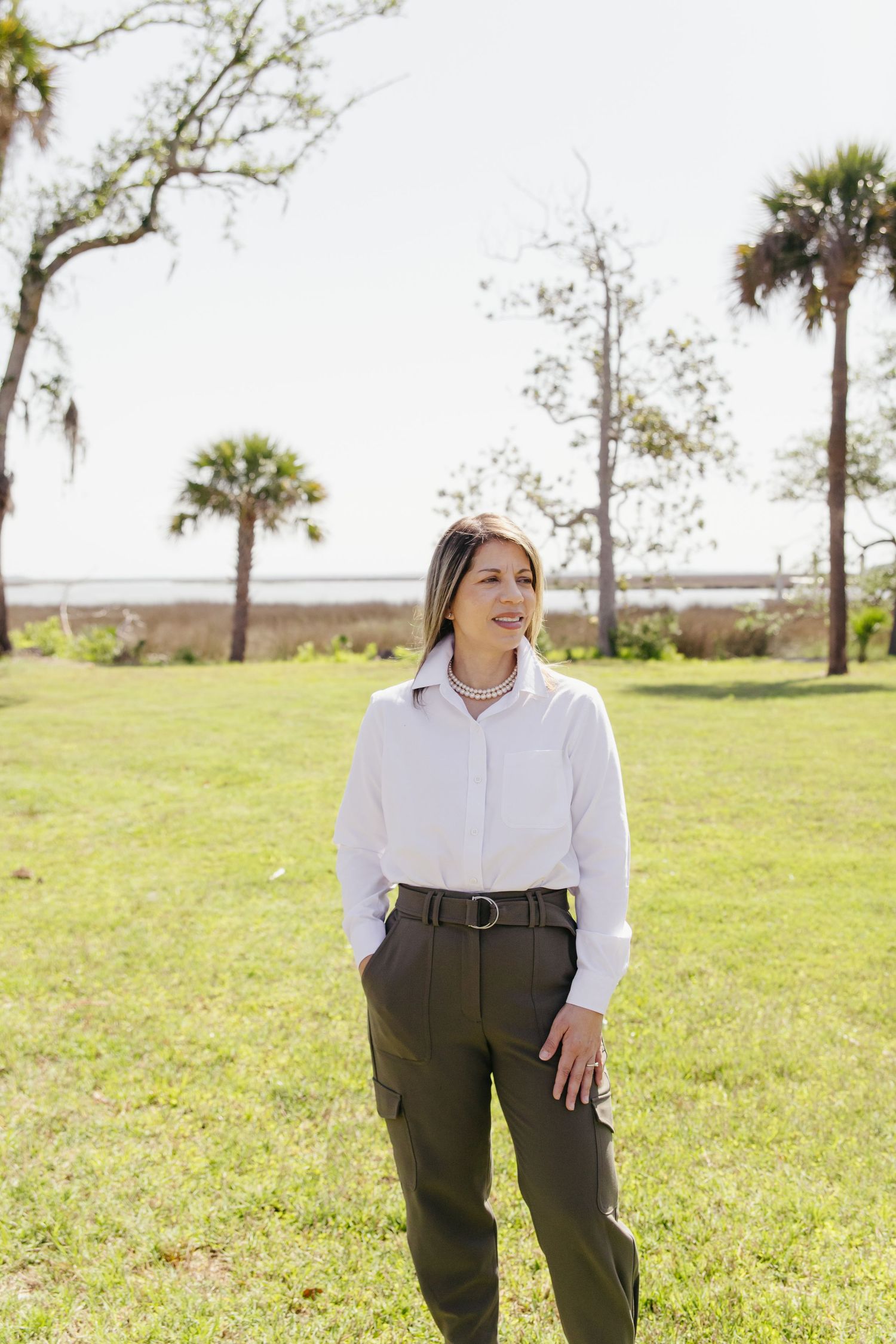 Professional woman in white shirt and olive pants standing on green lawn with palm trees behind her.