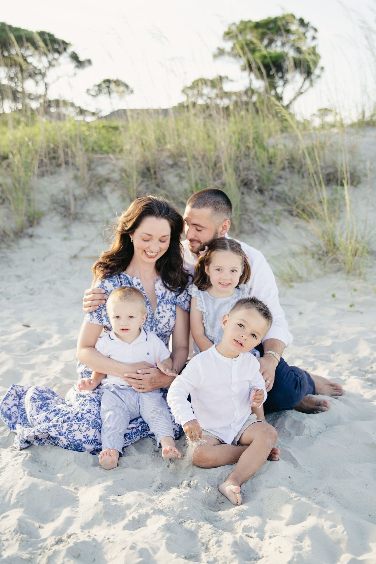 Happy family of five sits together on sandy beach, mother in floral dress with father and three young children.