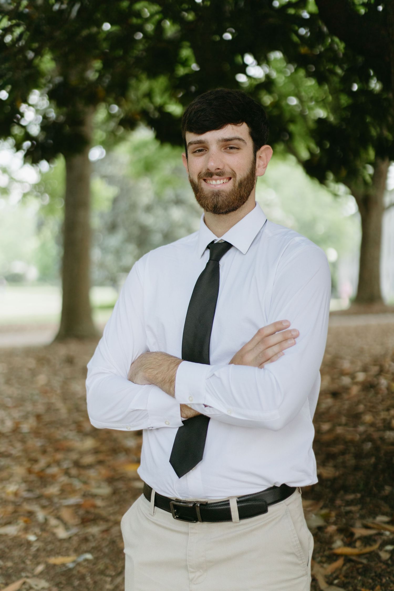 Young man with beard in white dress shirt and black tie stands with arms crossed outdoors among trees.