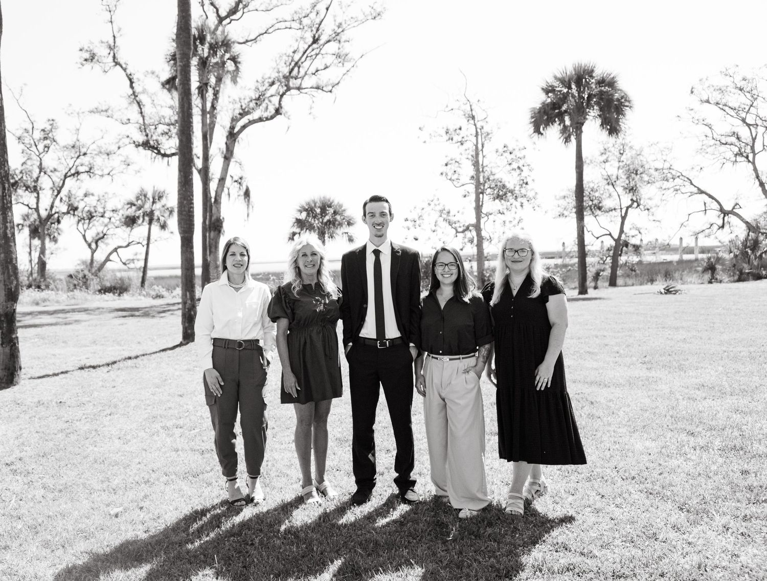 Professional business team of five posing outdoors among palm trees in black and white group photo.