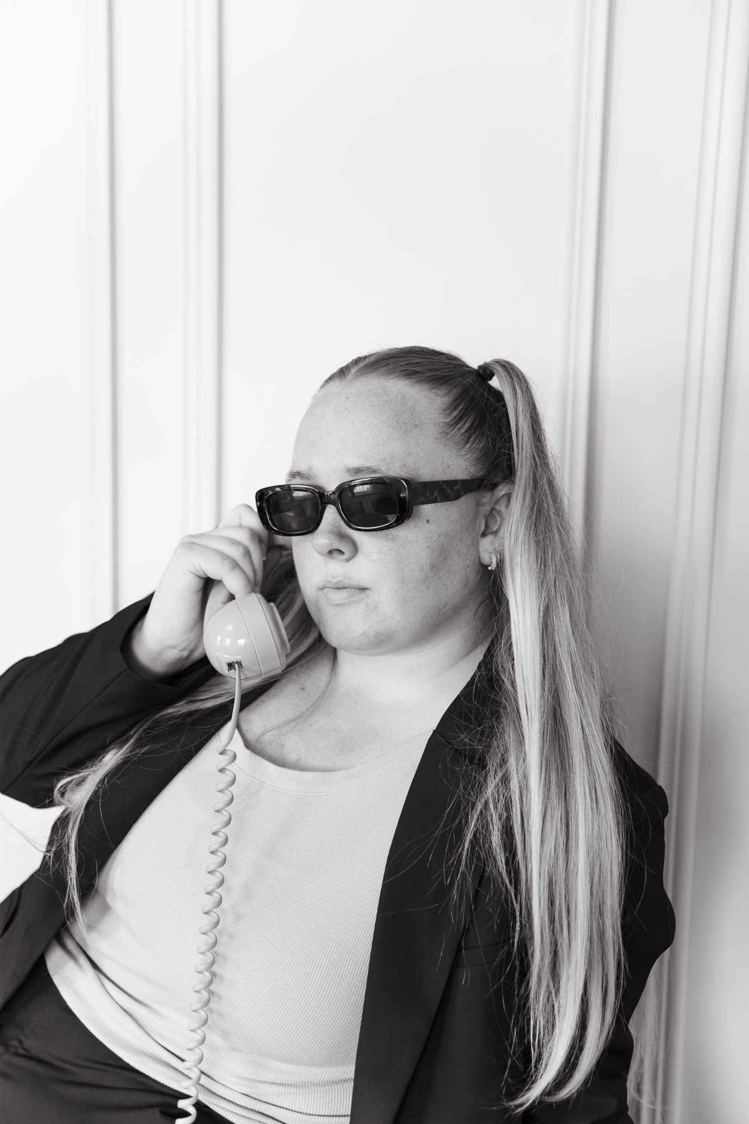 Professional woman in black blazer and sunglasses talking on vintage corded phone in black and white portrait.
