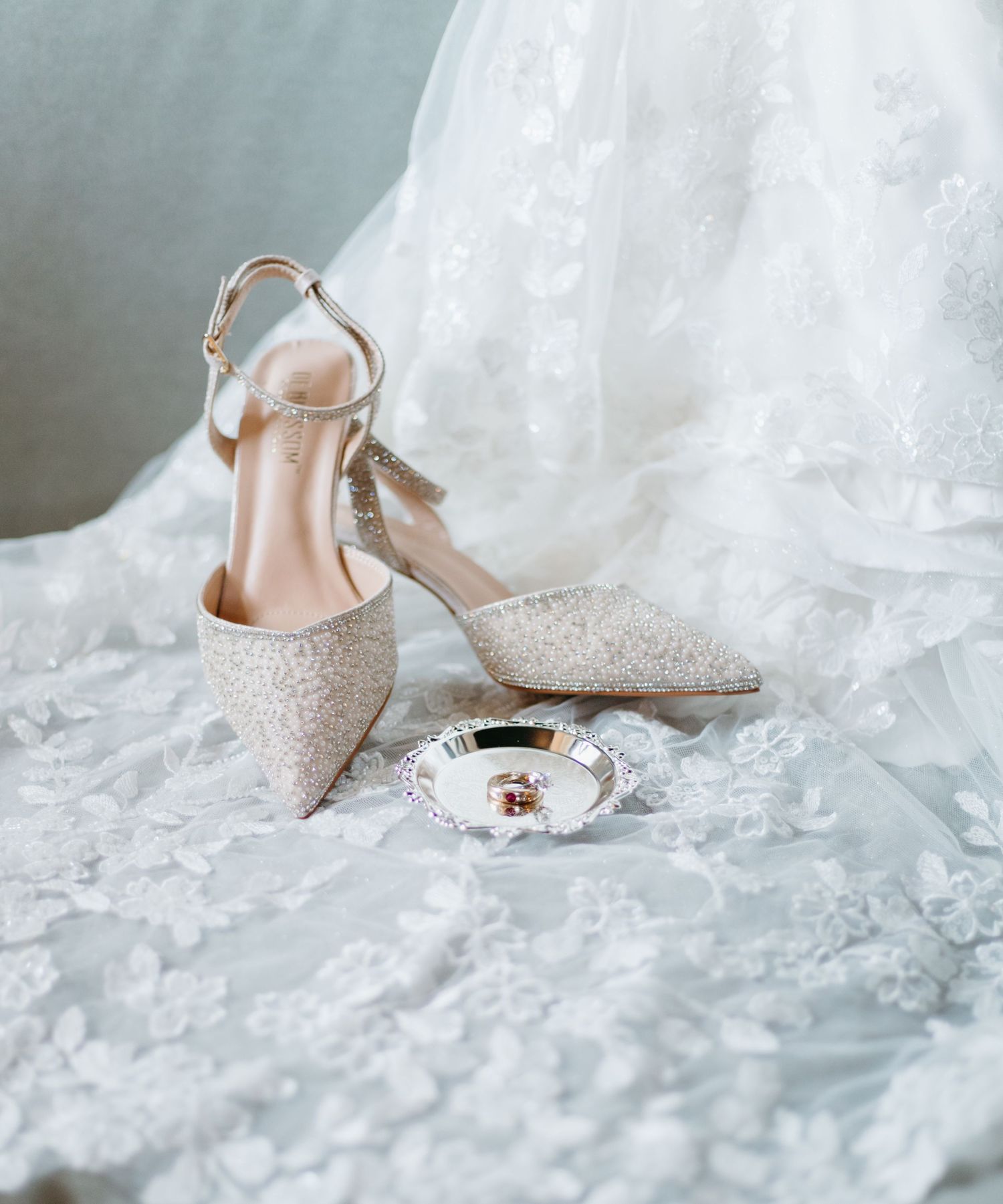 Glittery bridal heels with ankle straps displayed on lace wedding dress with small jewelry dish.