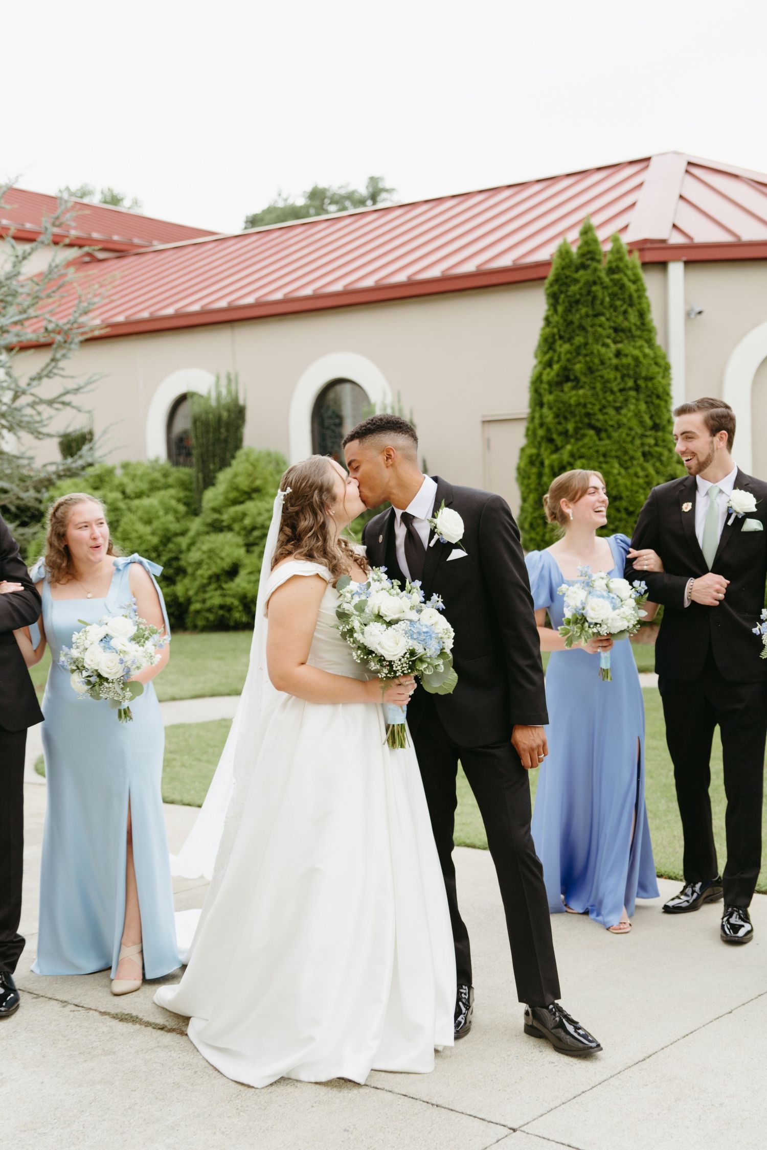 Newlywed couple kisses outside church surrounded by wedding party in blue dresses and black tuxedos.