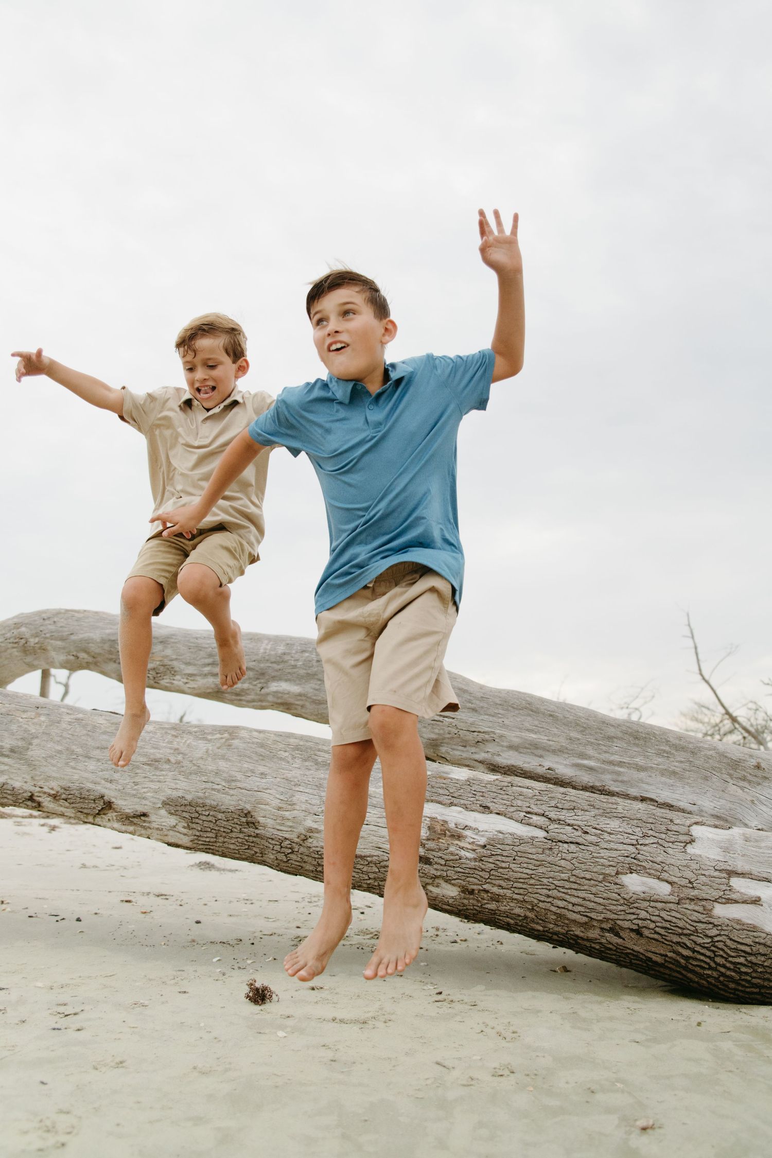 Two boys jump off a large driftwood log onto sandy beach, arms raised.