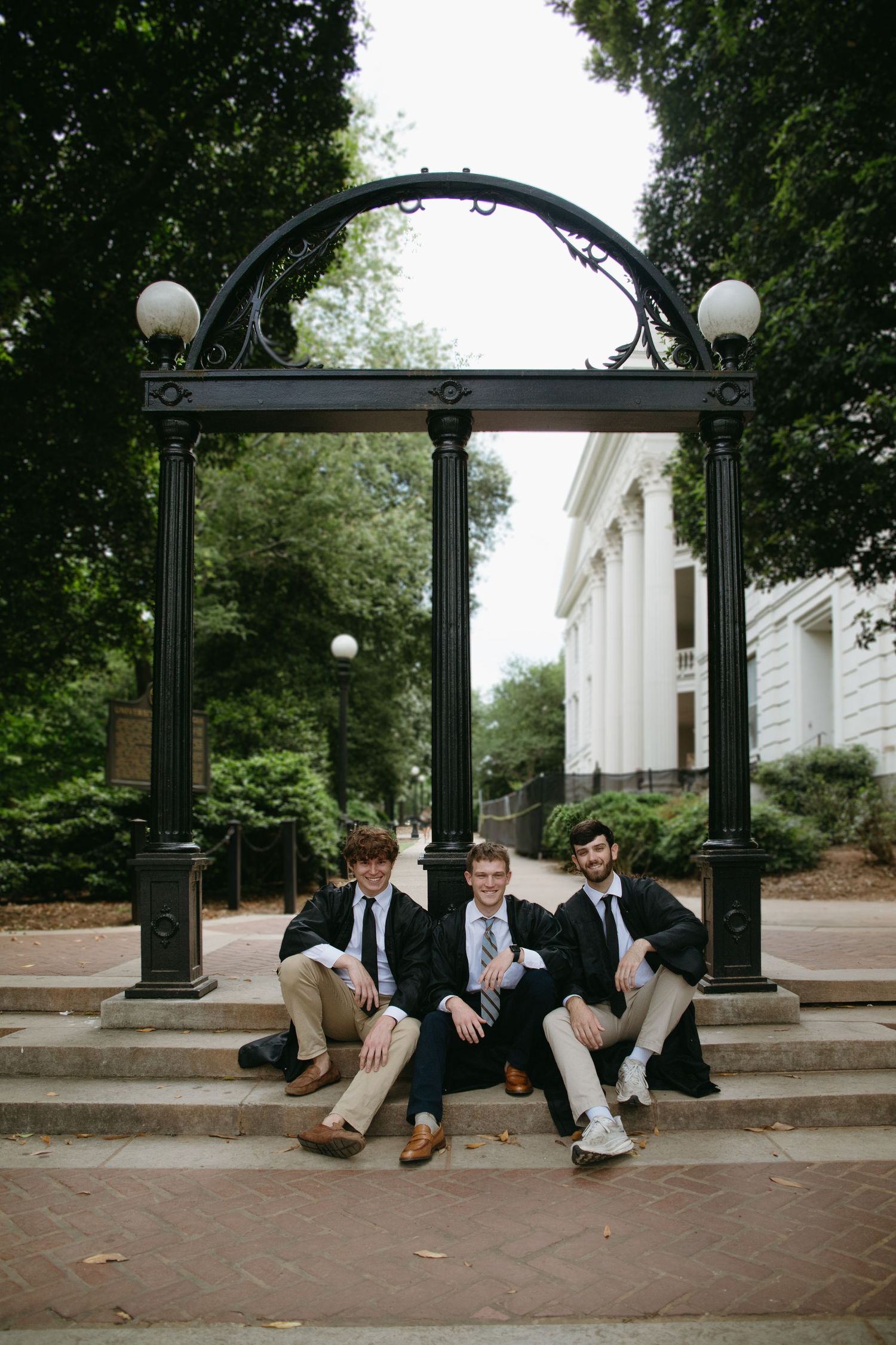 Three male graduates in blazers posing under iconic black arch on university campus for graduation portraits.