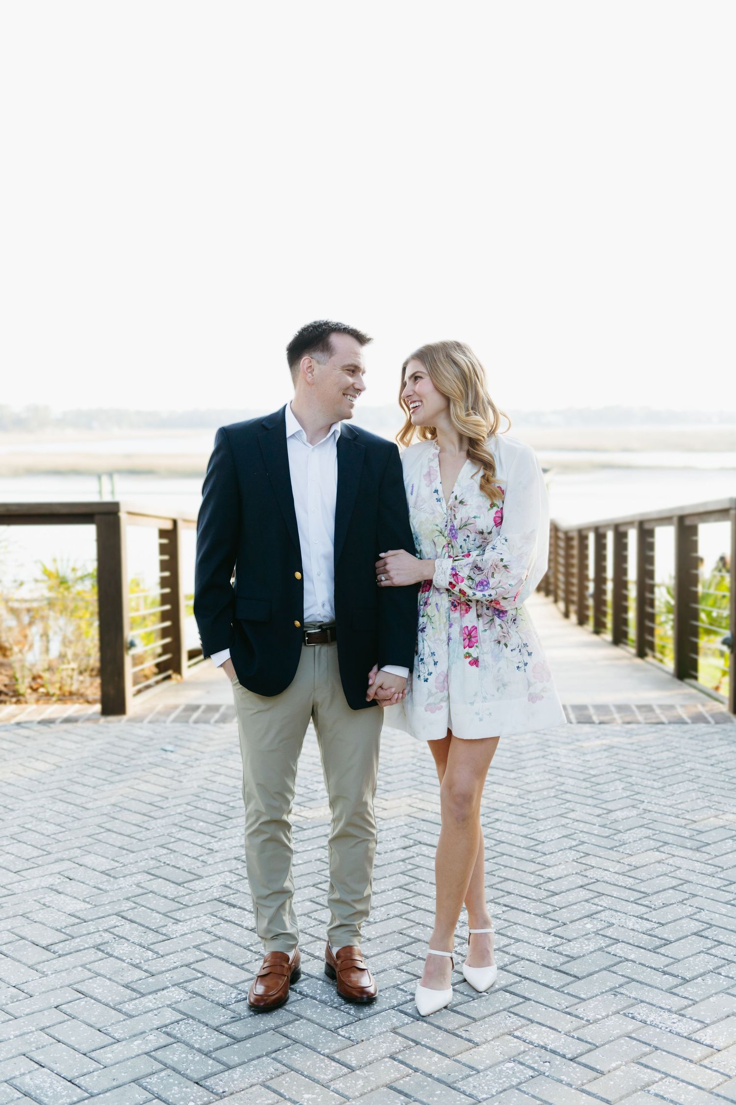Smiling couple holding hands on a waterfront boardwalk, woman in floral romper and man in navy blazer.