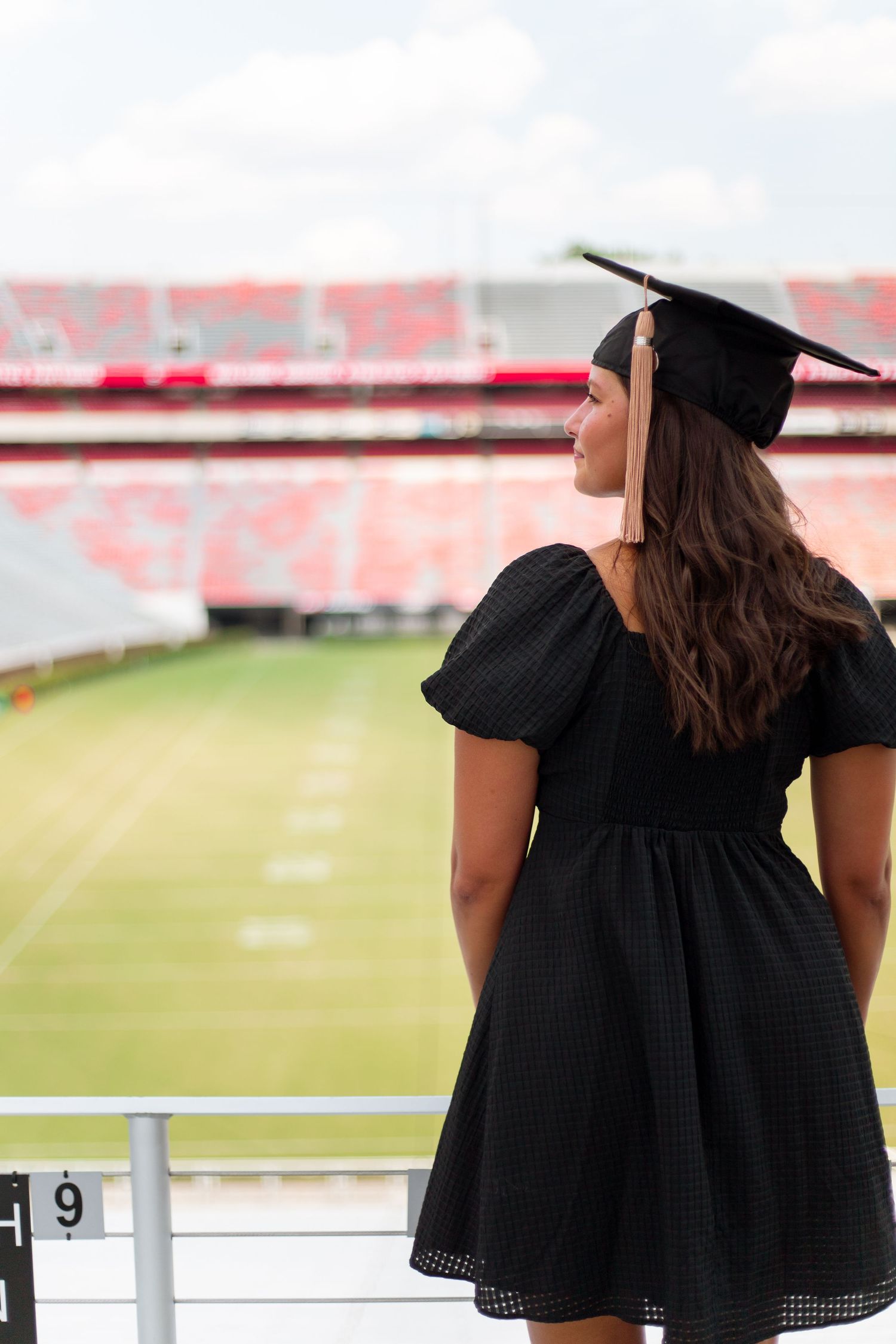 Female graduate in black dress and mortarboard cap gazing over football stadium field from stands.
