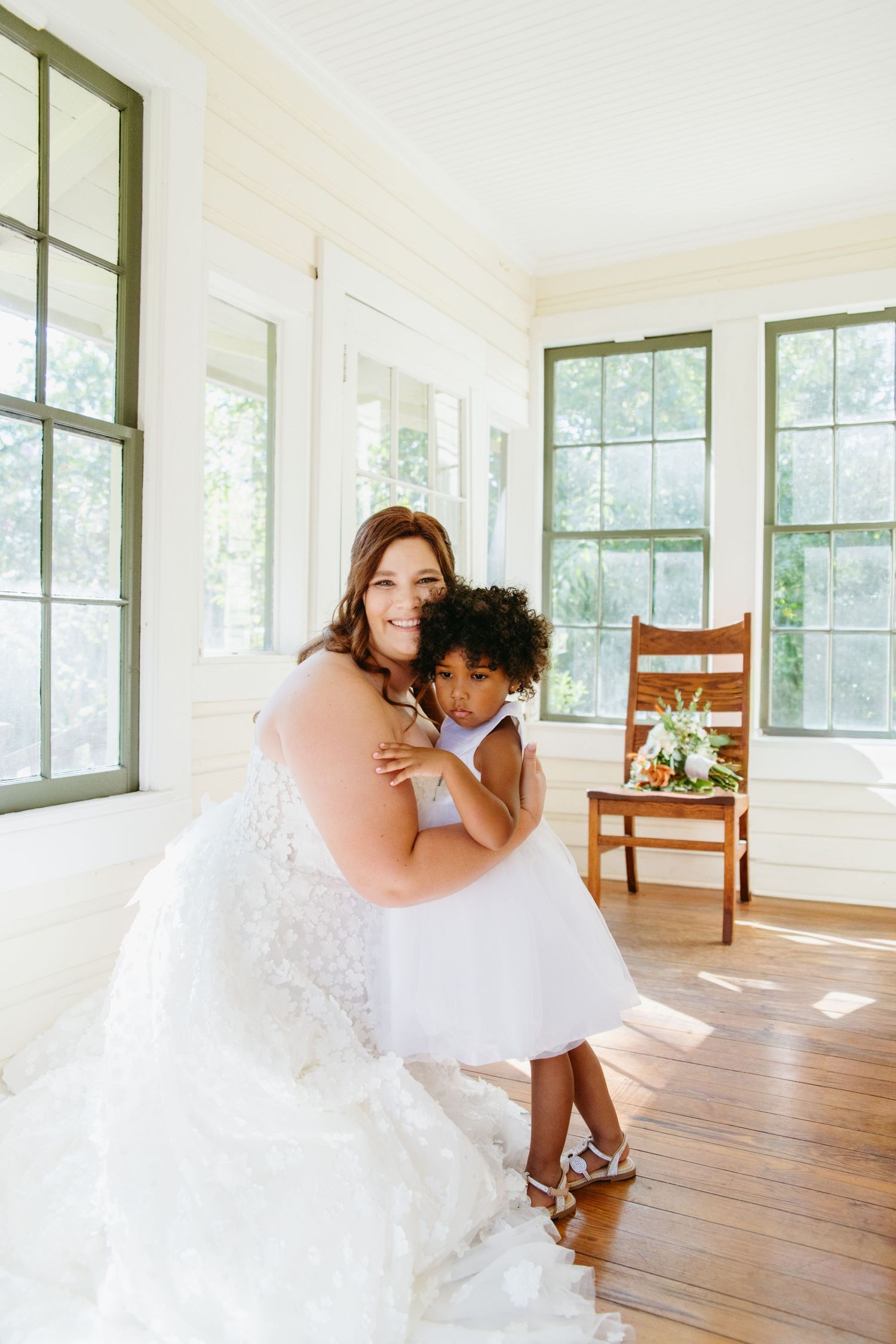 Smiling bride in lace gown kneels to embrace young flower girl in white dress in bright sunroom.