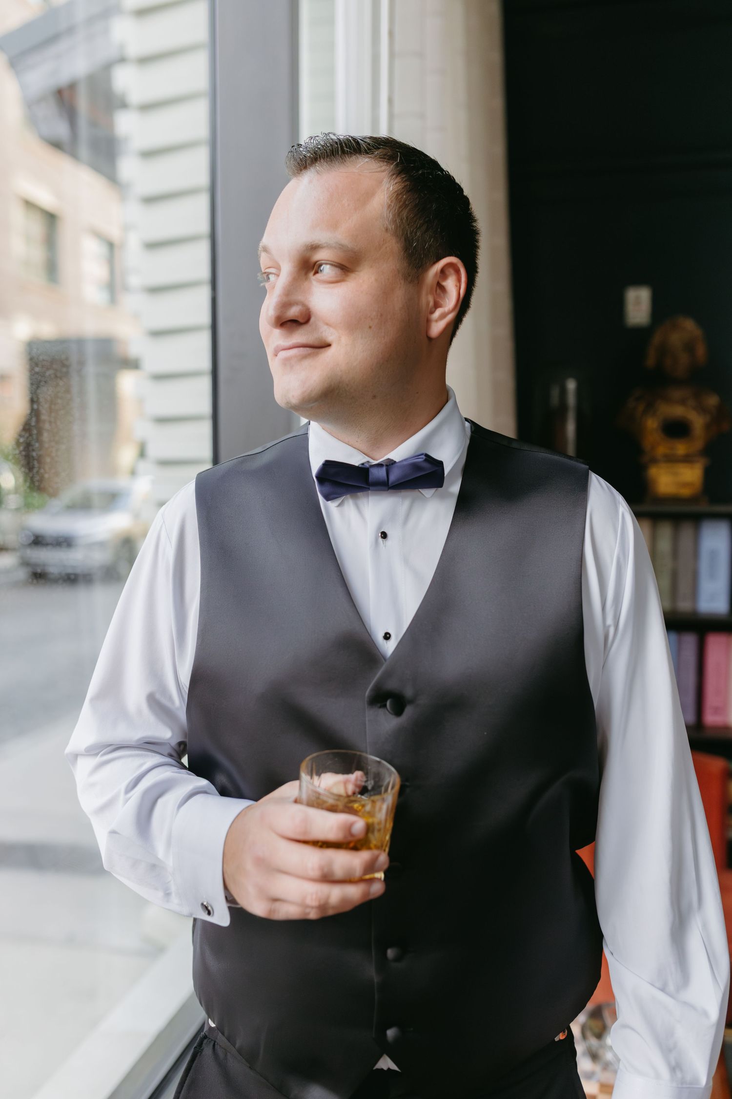Groom in gray vest and navy bow tie holding whiskey glass near window before wedding.
