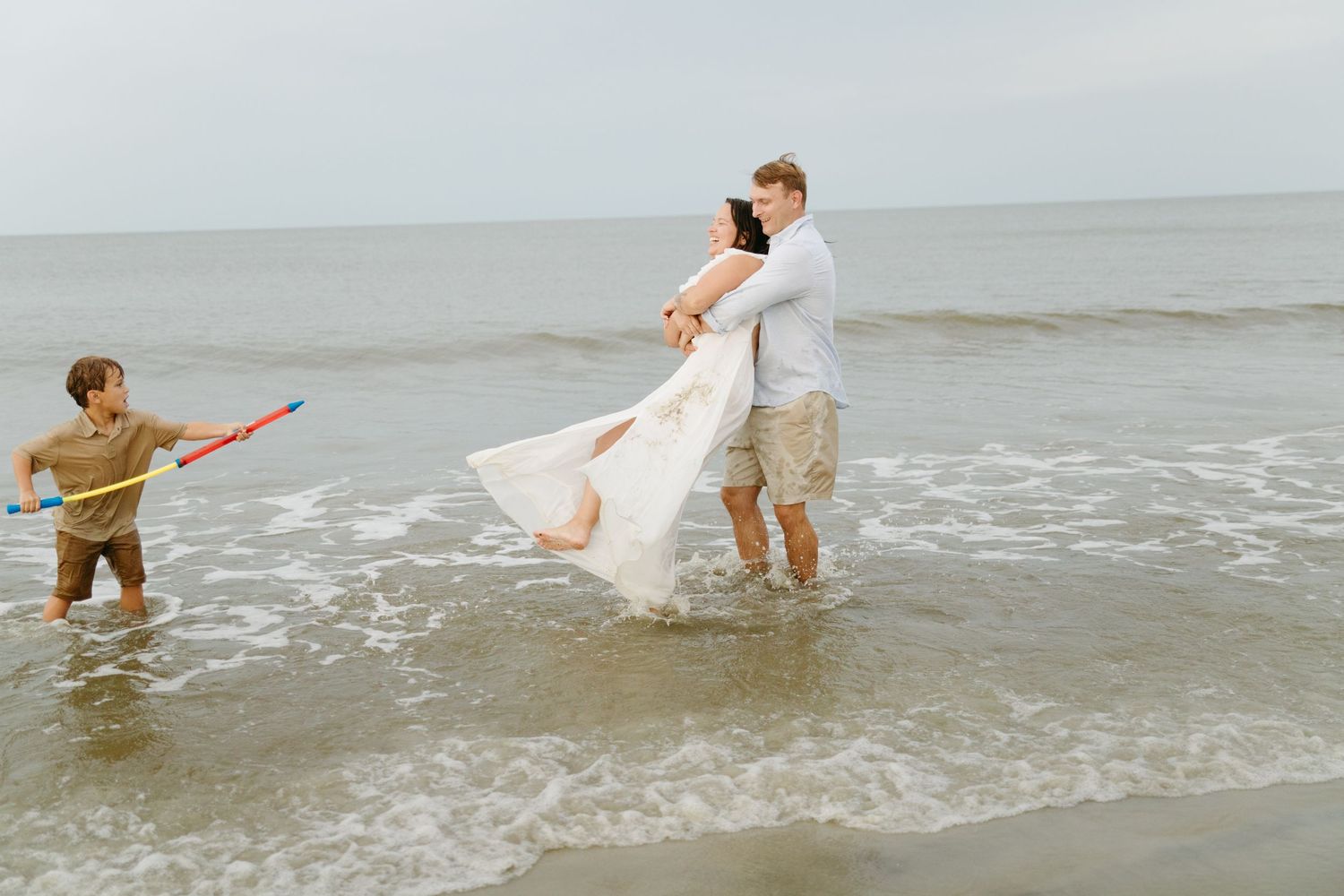 Couple embraces in ocean waves while a child plays nearby with a toy.