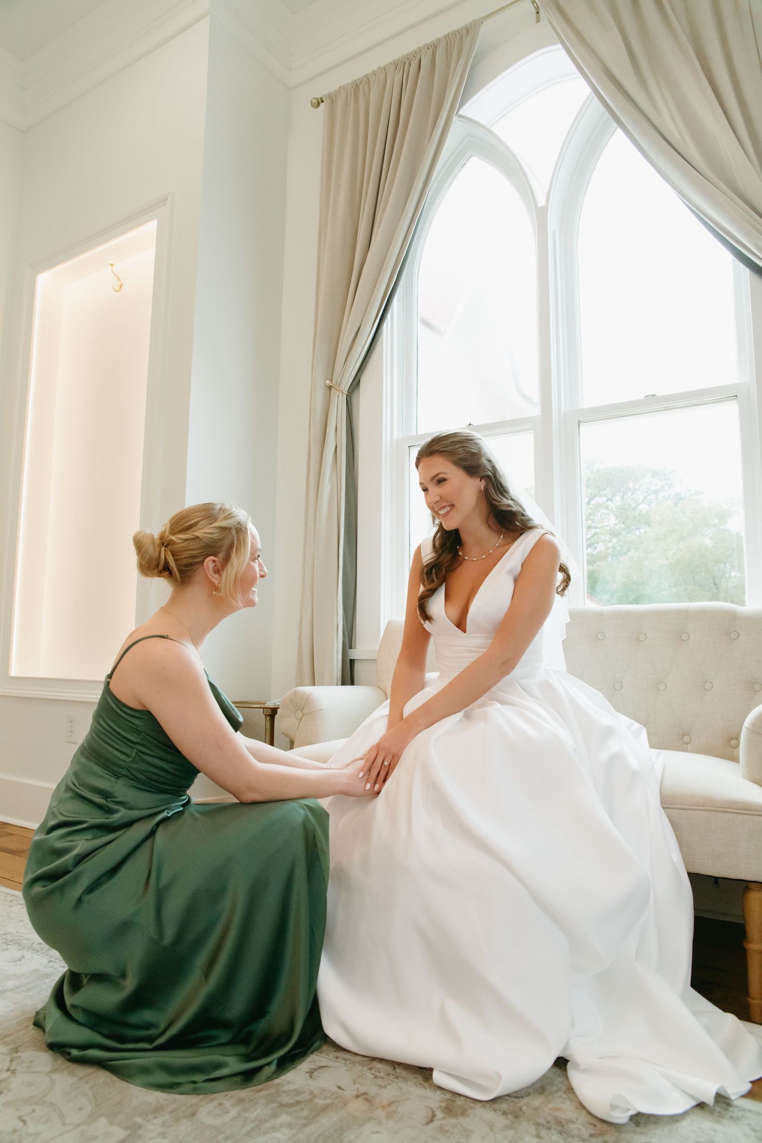 Bridesmaid in green dress kneeling beside smiling bride in white gown by arched window.