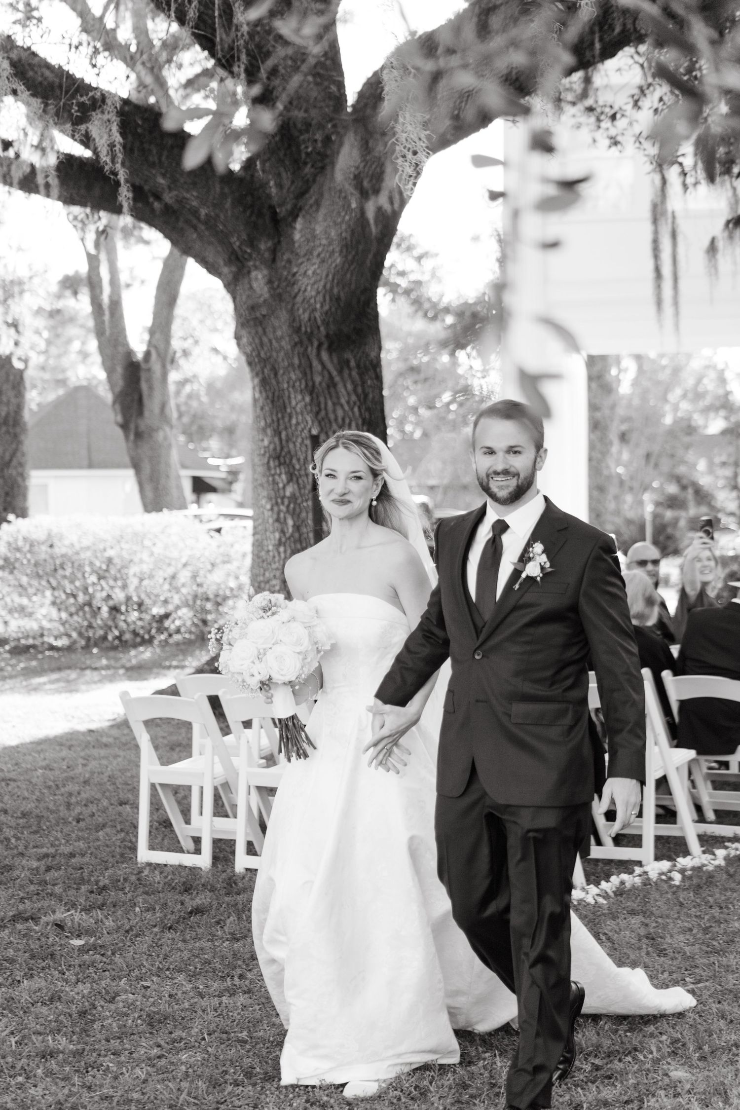 Newlyweds walk hand-in-hand down the aisle after ceremony beneath large oak tree in black and white.