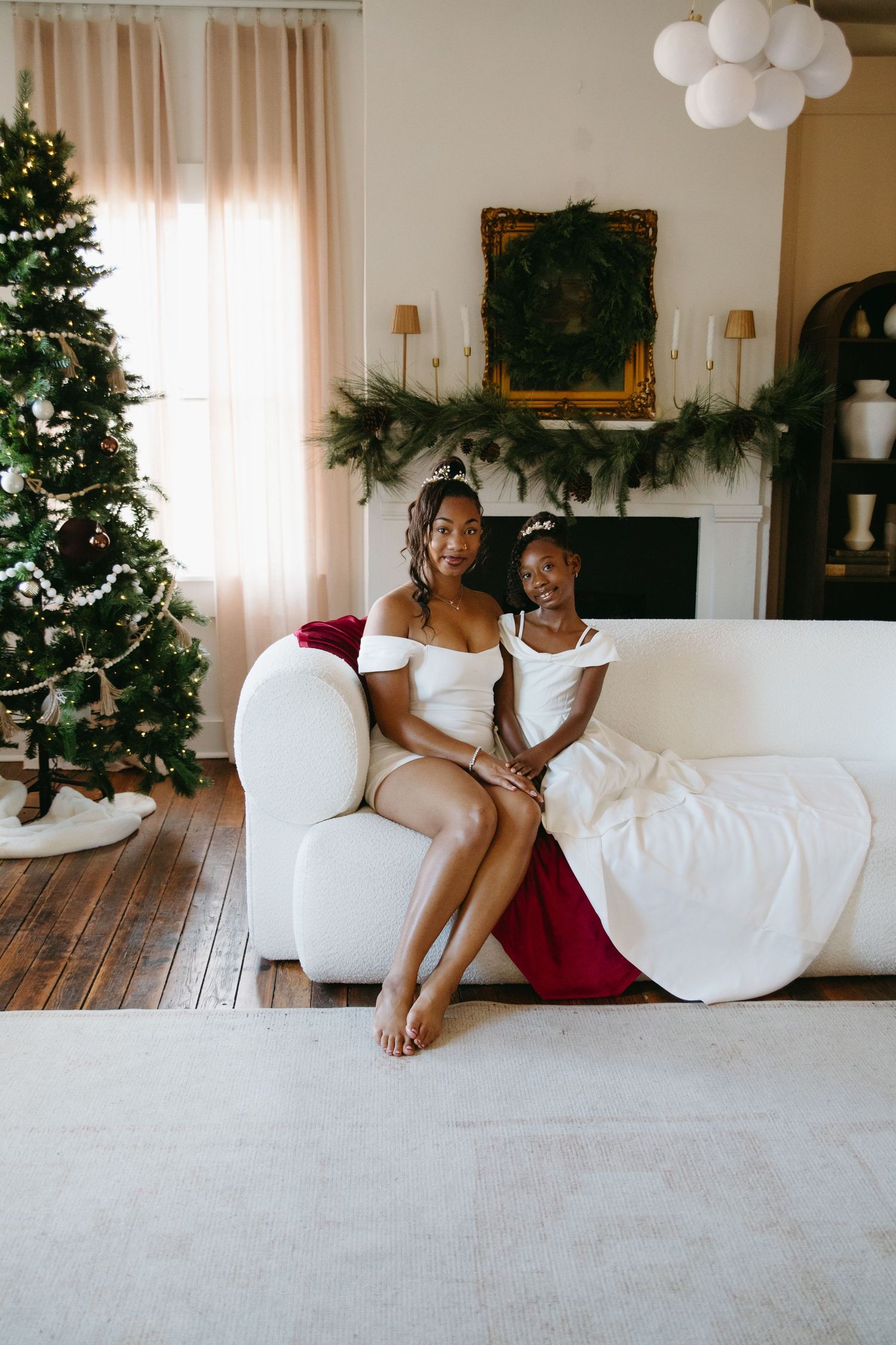 A mother and daughter in white dresses pose together on a white sofa with a decorated Christmas tree and garland fireplace behind them.