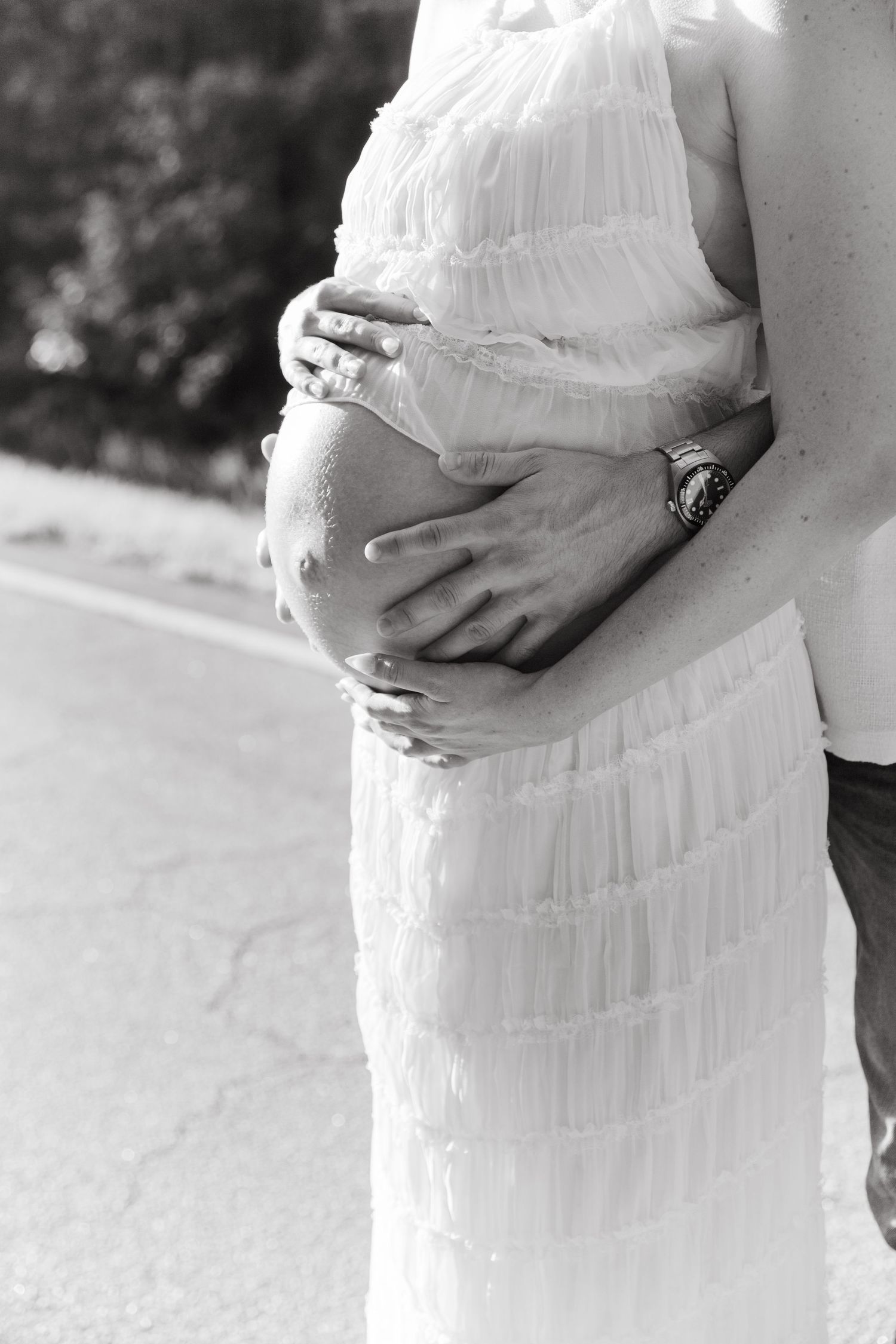 Black and white photo of a pregnant woman in white dress with partner's hands cradling her baby bump.