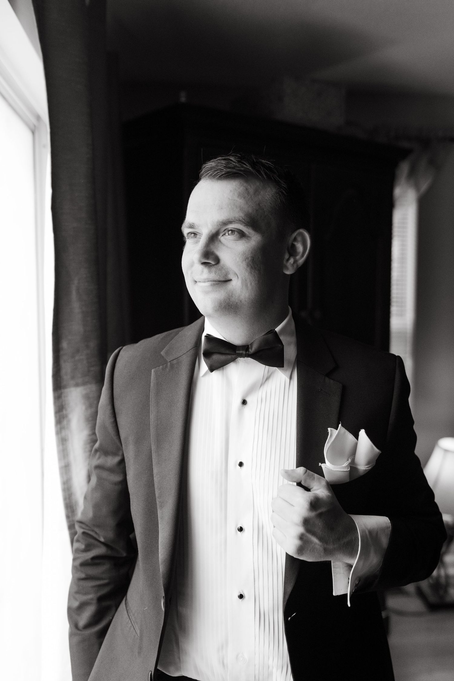 Black and white portrait of a groom in a tuxedo with bow tie gazing out a window.