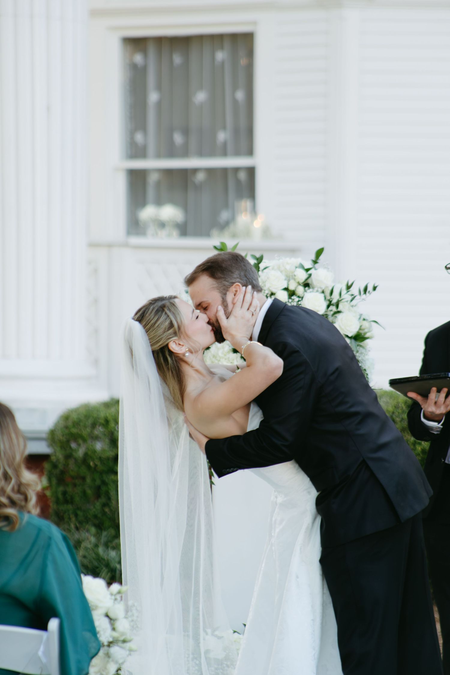 Bride and groom share their first kiss surrounded by white floral arrangements at outdoor ceremony.