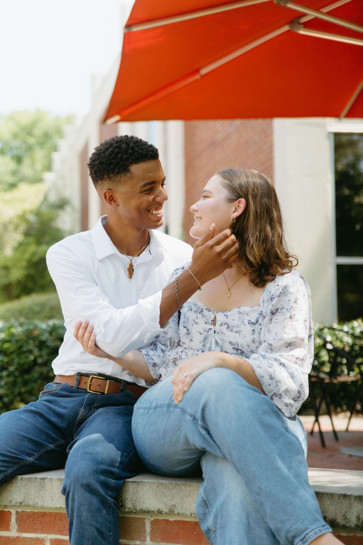 Smiling interracial couple sitting outdoors on a brick wall under a red umbrella, man tenderly touching woman's face.