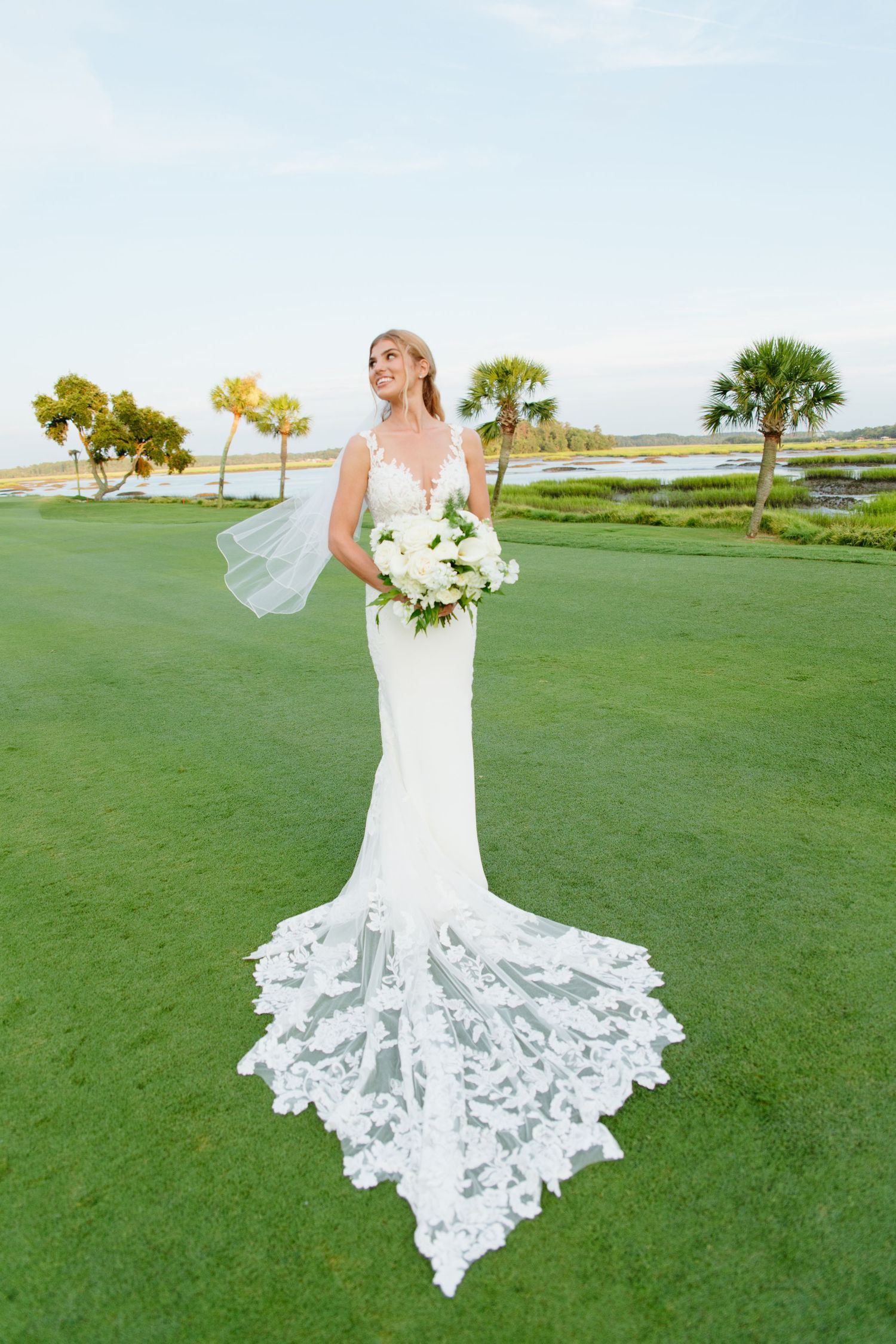 Elegant bride in a fitted lace wedding gown with cathedral train holds white bouquet on a golf course near water.