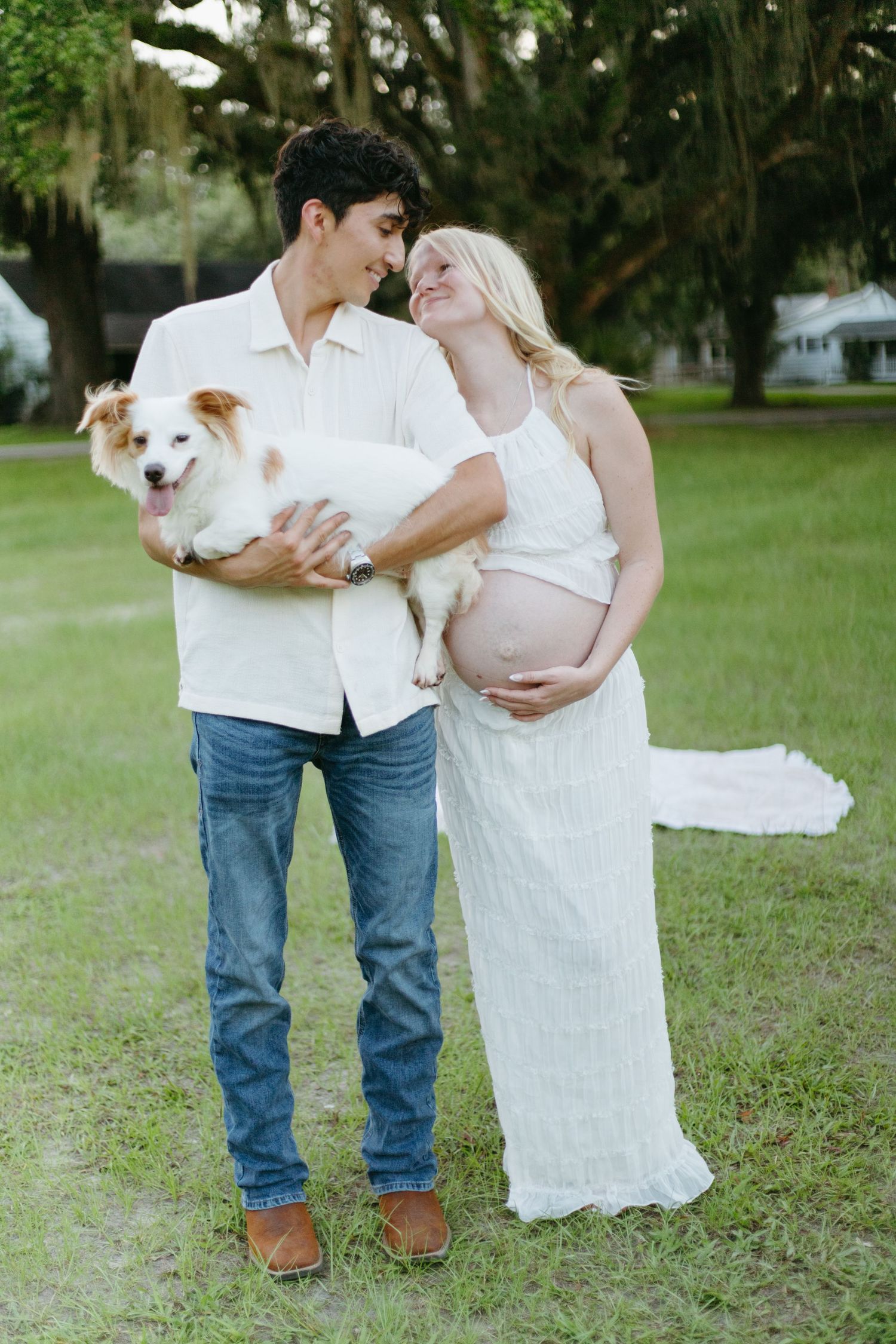 Happy expecting couple holding a white dog while standing together in a green park.