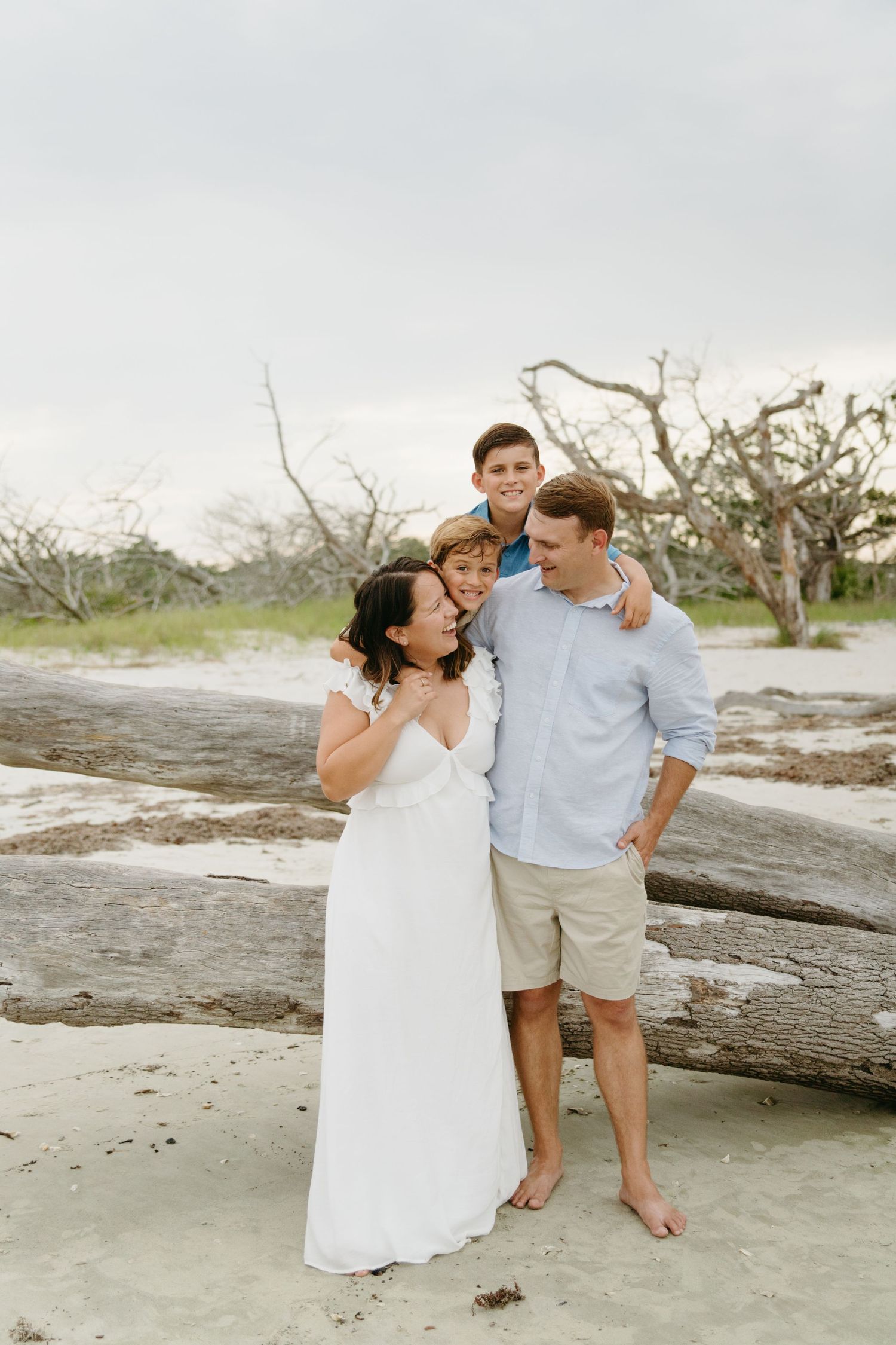 A happy family of four poses casually on a beach with driftwood and trees in the background.