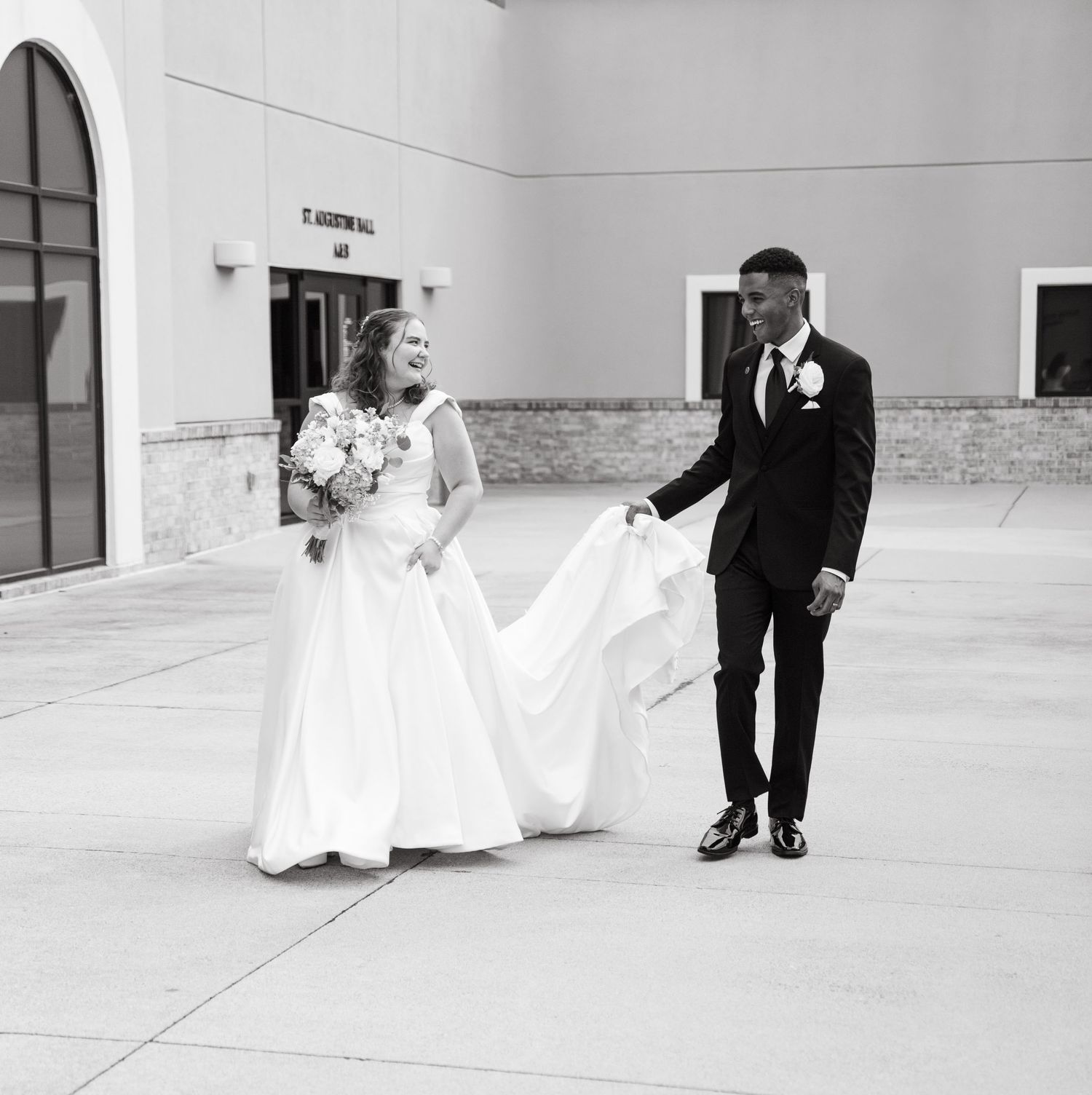 Groom helps bride with wedding dress train in black and white photo outside St. Andrew Hall.