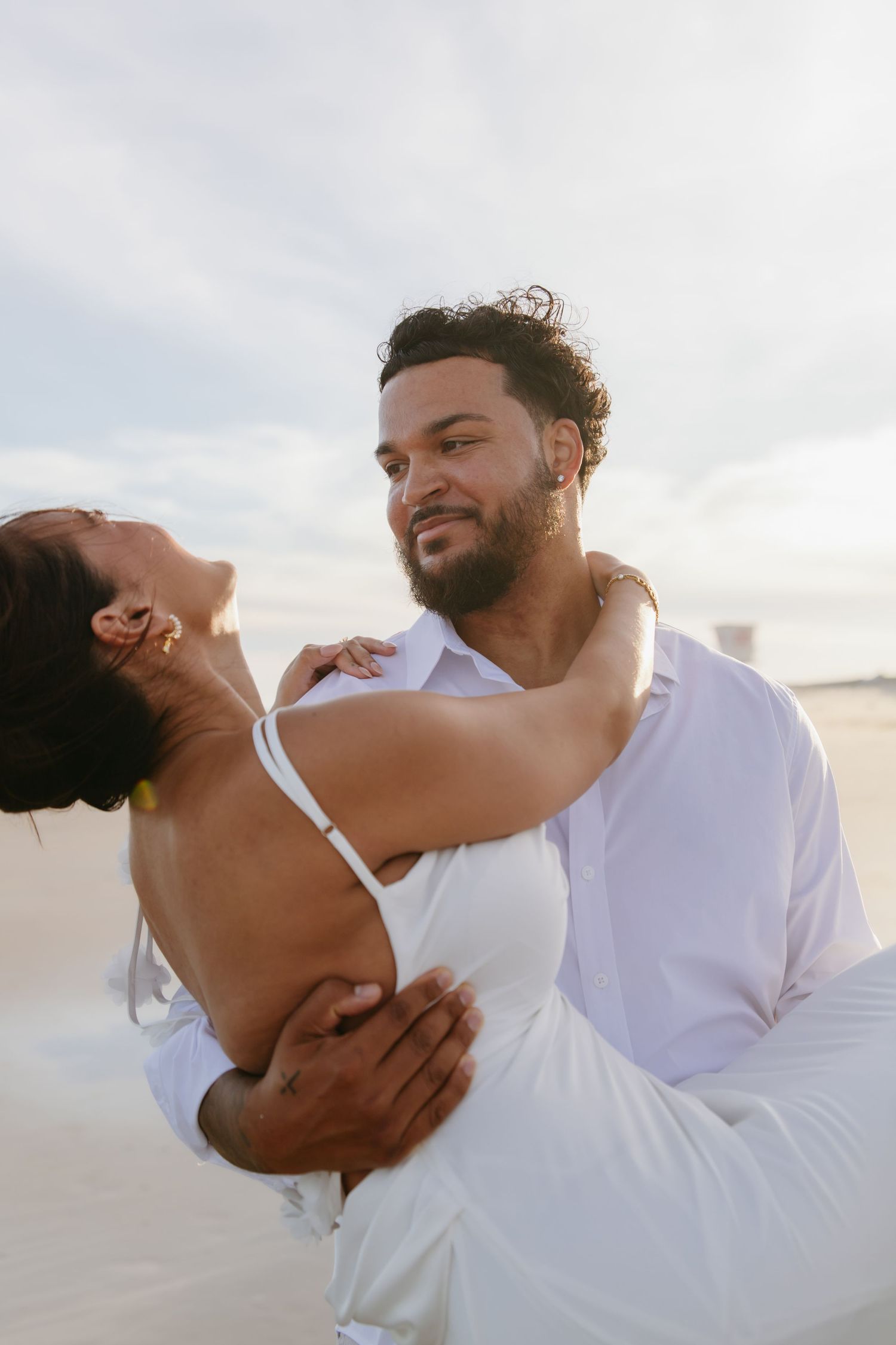Romantic couple embracing on a sandy beach, man in white shirt dipping woman in white dress.