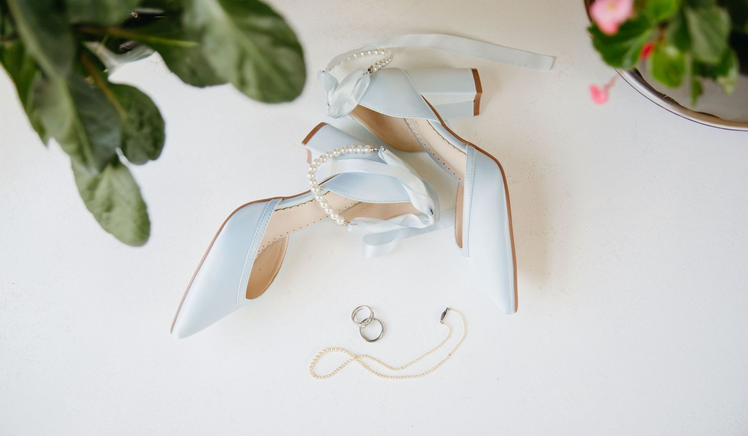 Light blue strappy heels with wedding rings on white surface near plant.