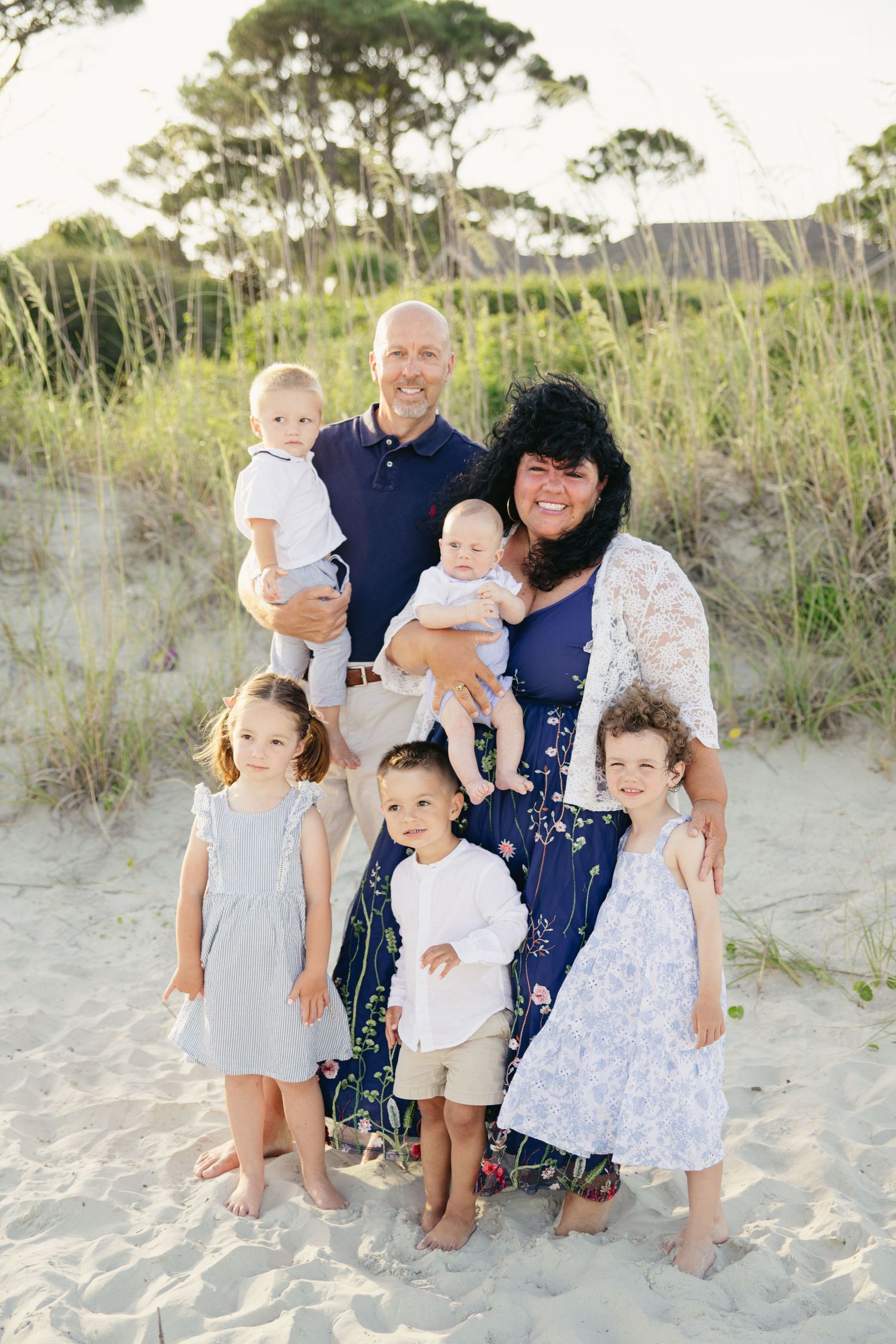 Family of seven poses on sandy beach dunes with lush greenery, parents holding two babies with three children standing in front.