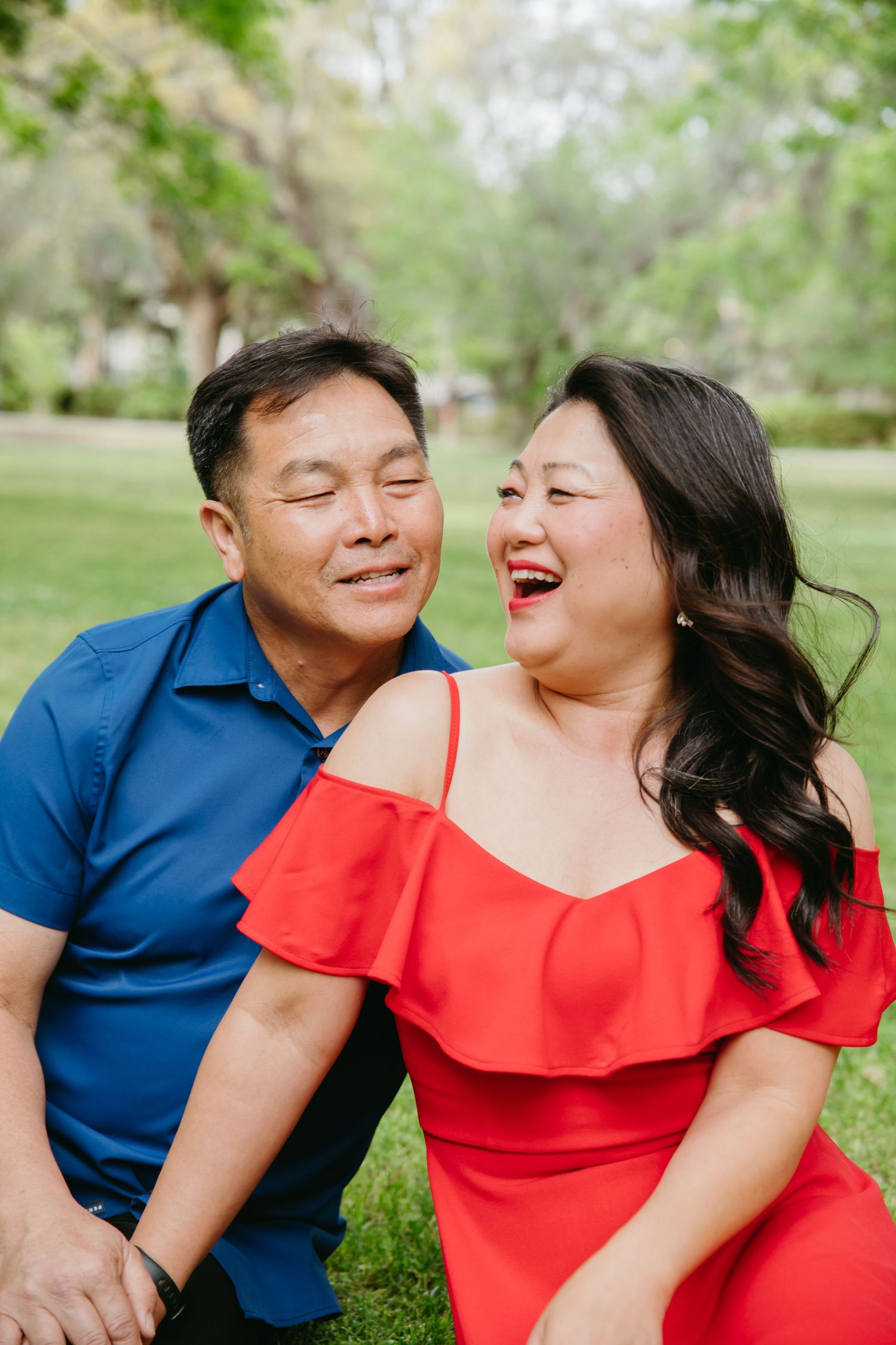 Asian couple laughing joyfully together while sitting on green grass in a sunny park.