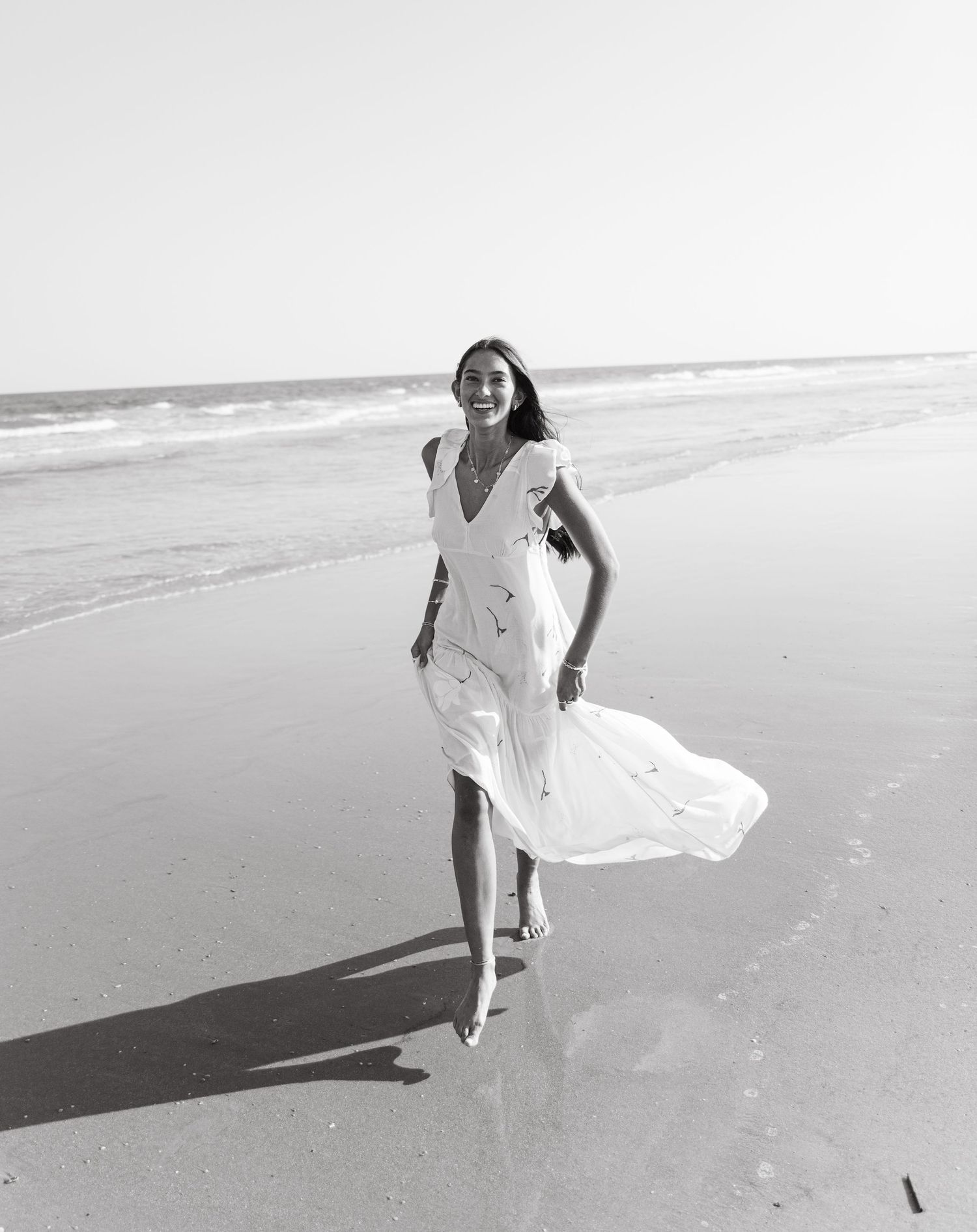 Black and white photo of a woman in a flowing white dress walking along a sunny beach shoreline.