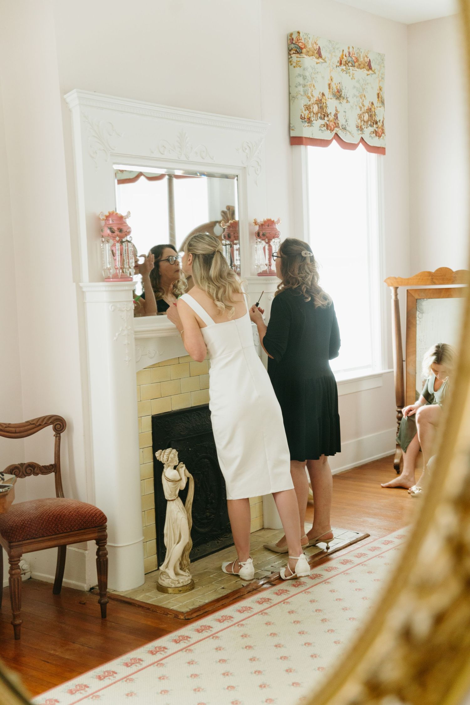 Bride in white dress stands before fireplace mirror with bridesmaid helping her prepare on wedding day.
