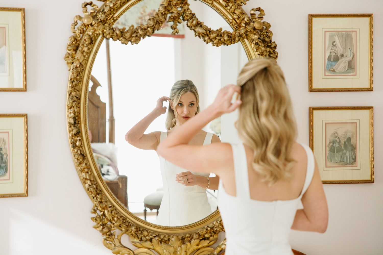 Bride adjusting her veil in an ornate gold mirror while wearing a white wedding gown.