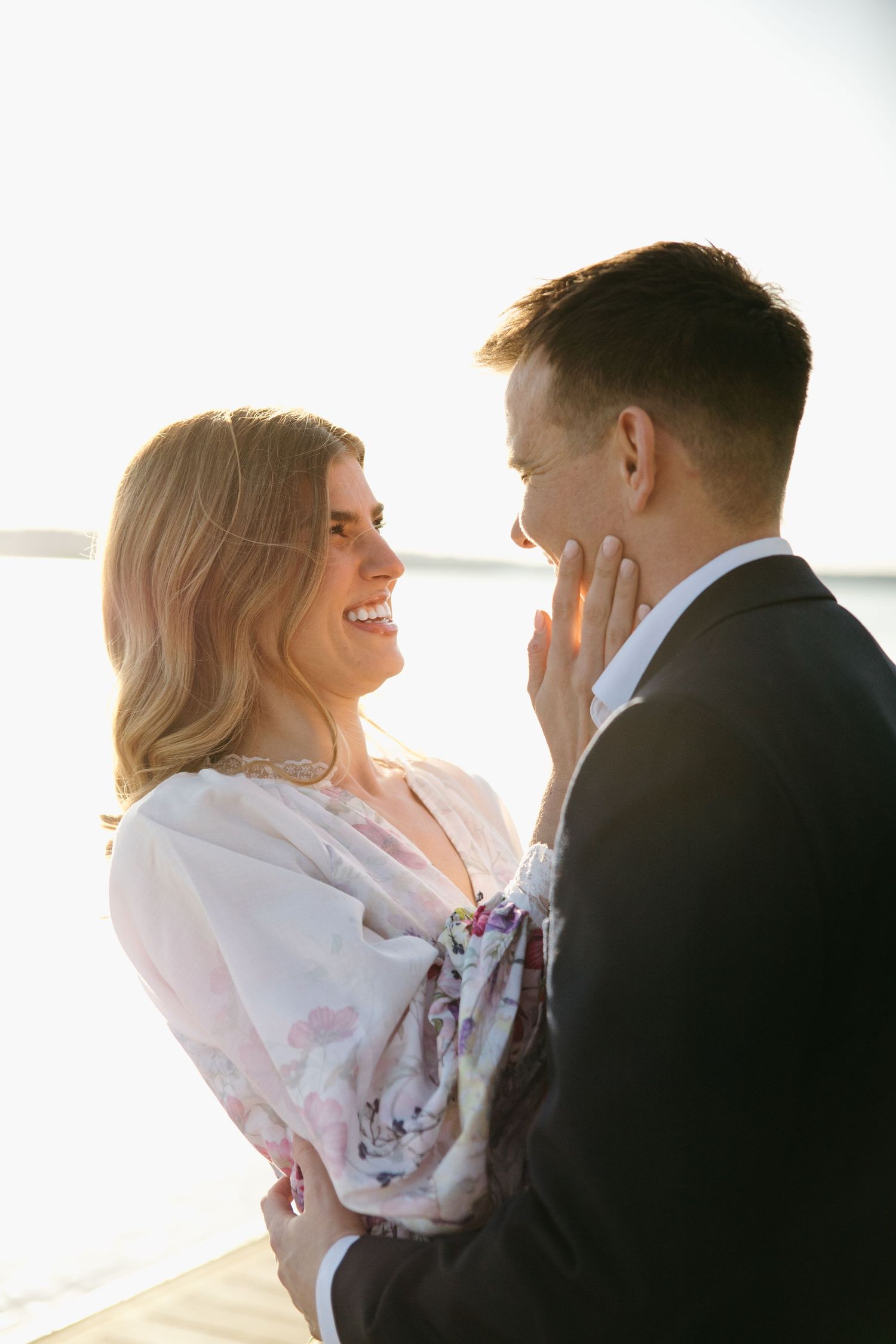 Couple sharing a romantic moment at sunset by the water, smiling and touching faces tenderly.