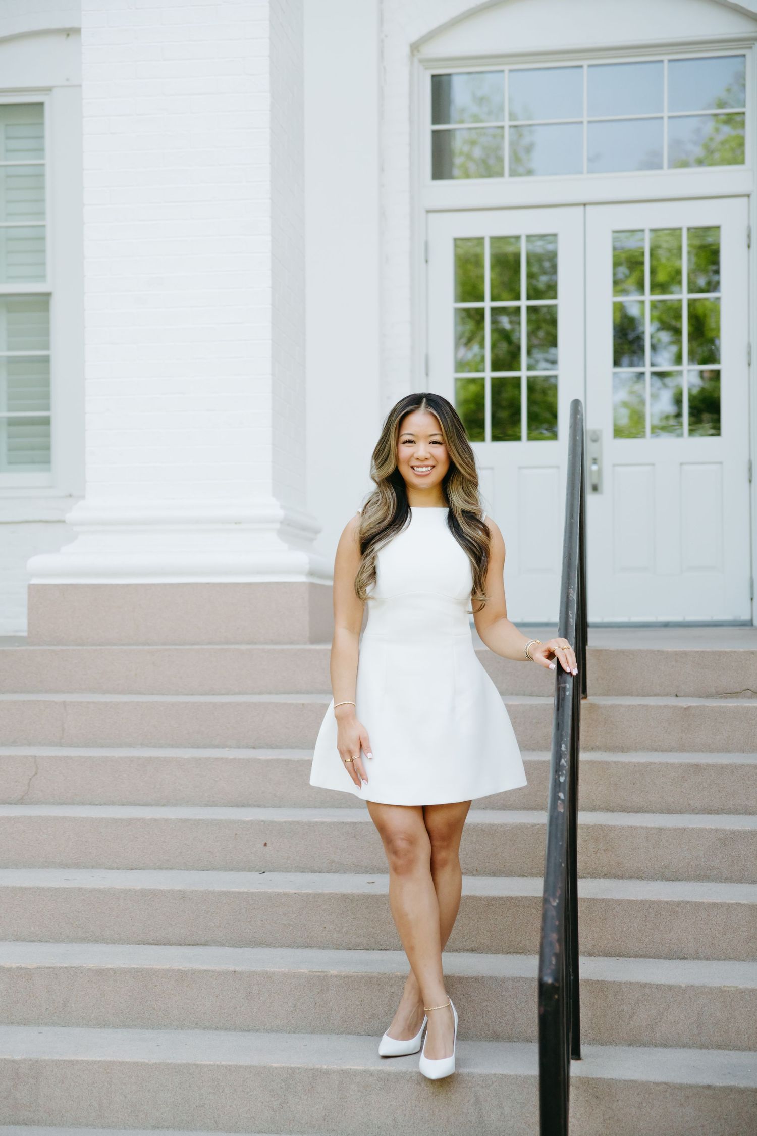 Asian woman in white sleeveless dress standing on steps of white building smiling.