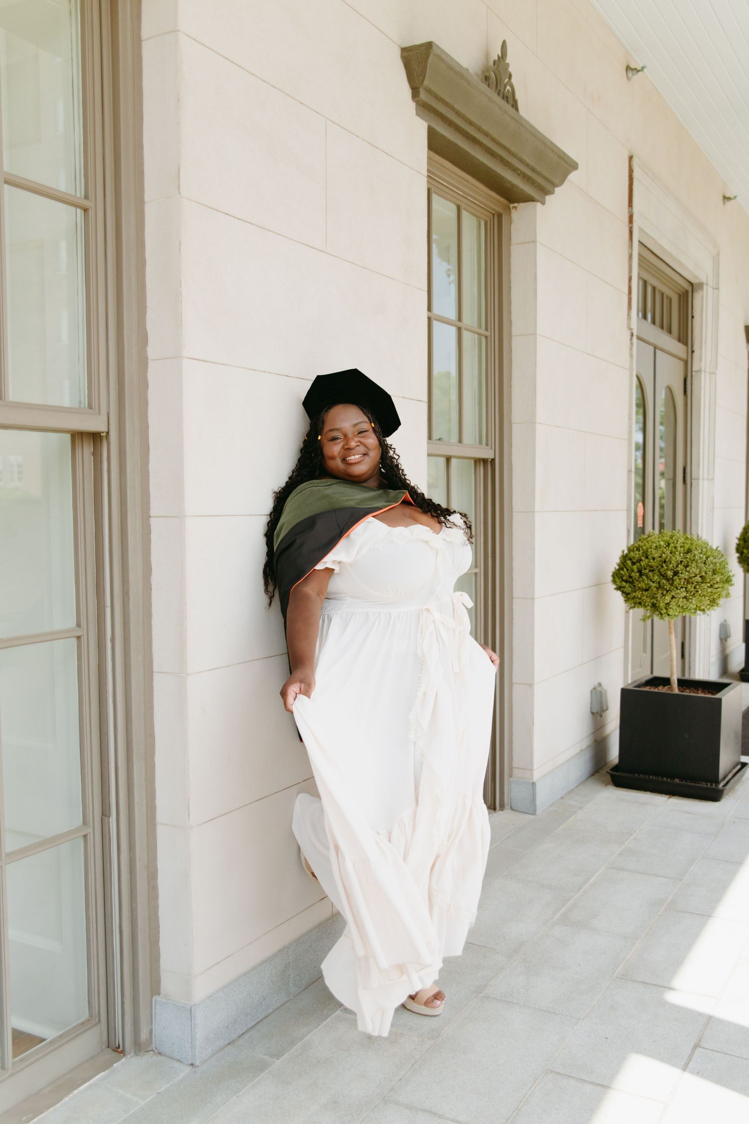 Black woman in white maxi dress and graduation cap leaning against elegant building.