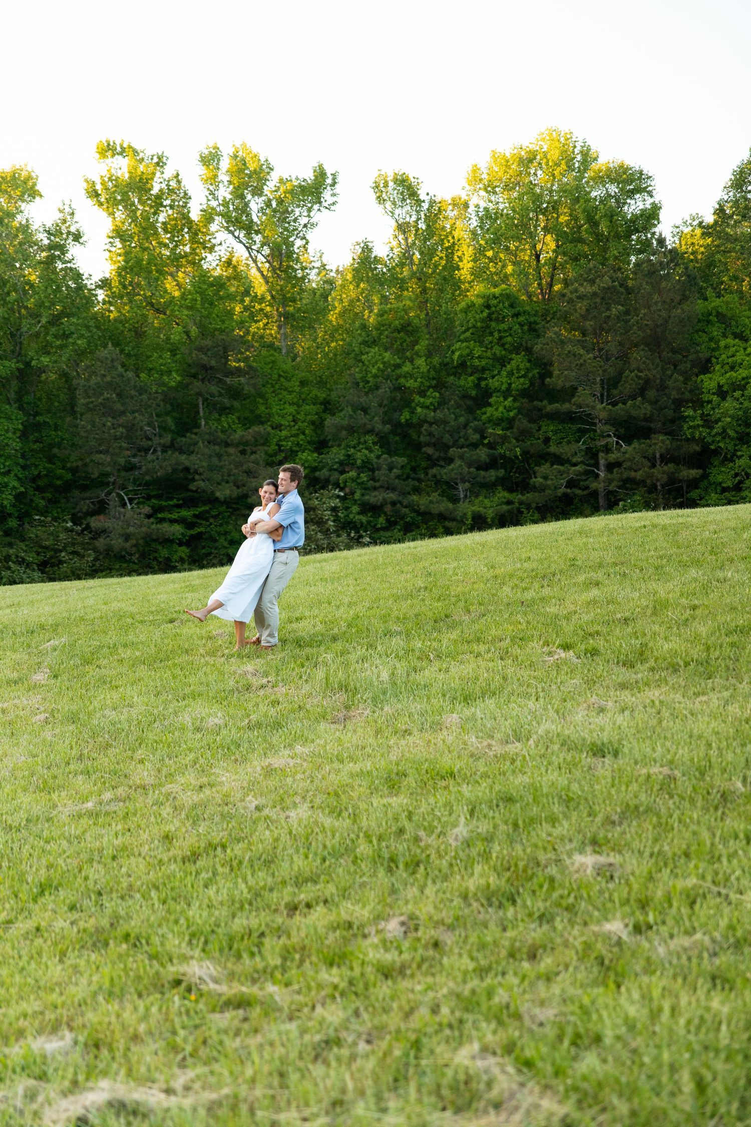 Couple embracing on a green hillside with tall trees in background during a golden hour engagement shoot.