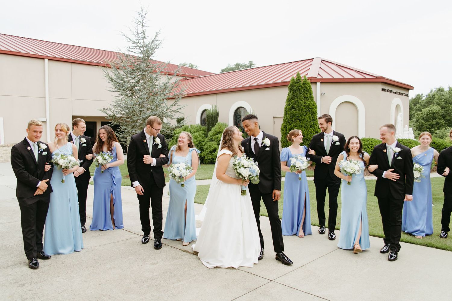 Wedding party in blue dresses and black tuxedos posing outside an elegant venue.