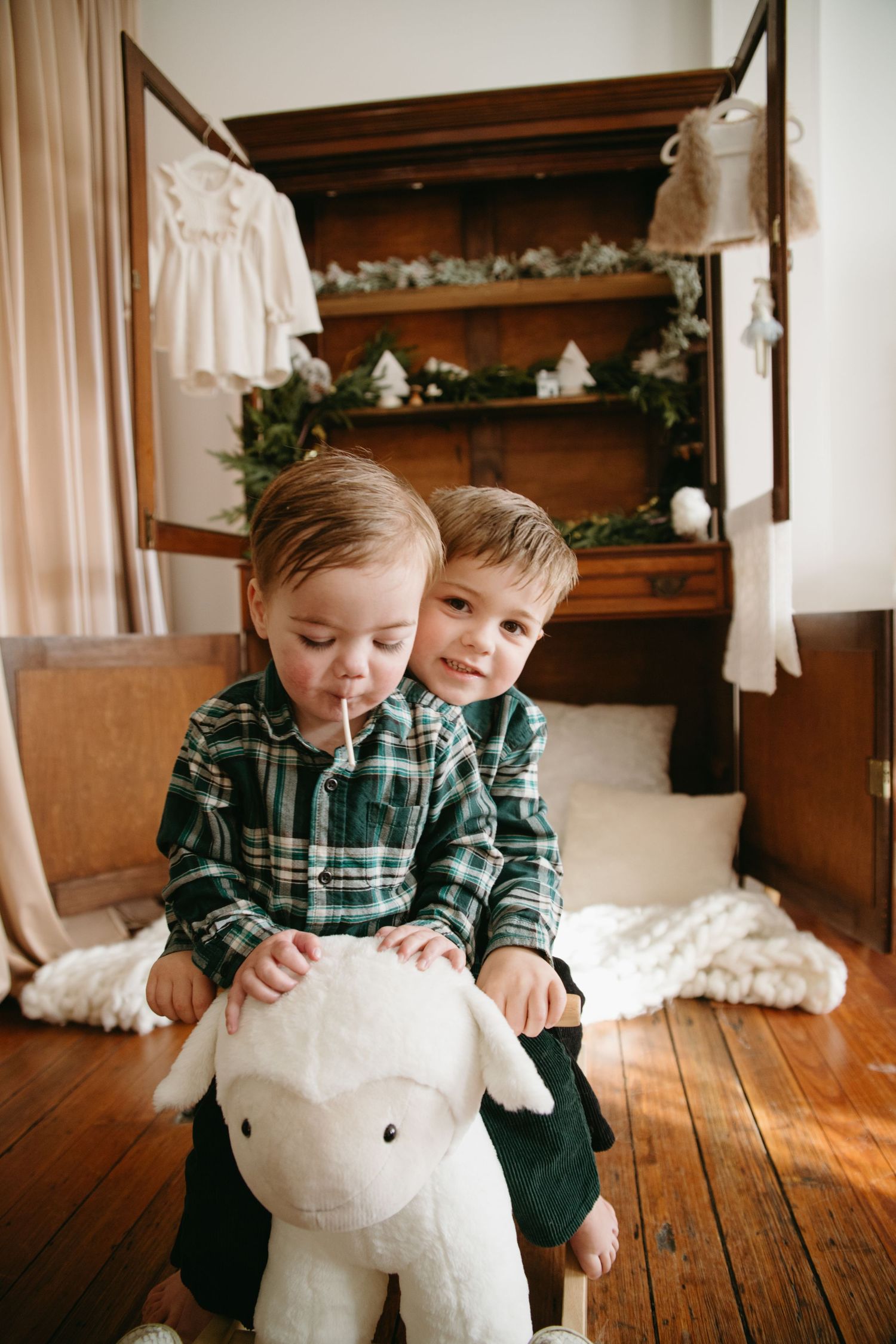 Two young brothers in matching green plaid shirts share a white lamb rocking toy in a rustic Christmas-decorated bedroom.