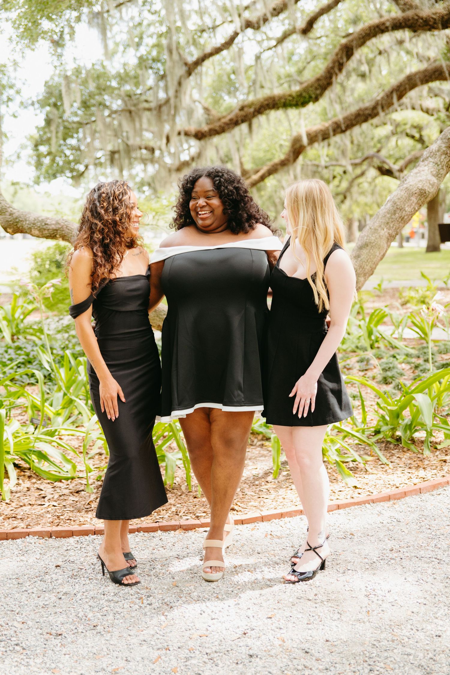 Three women in black dresses laughing together on a tree-lined park path.