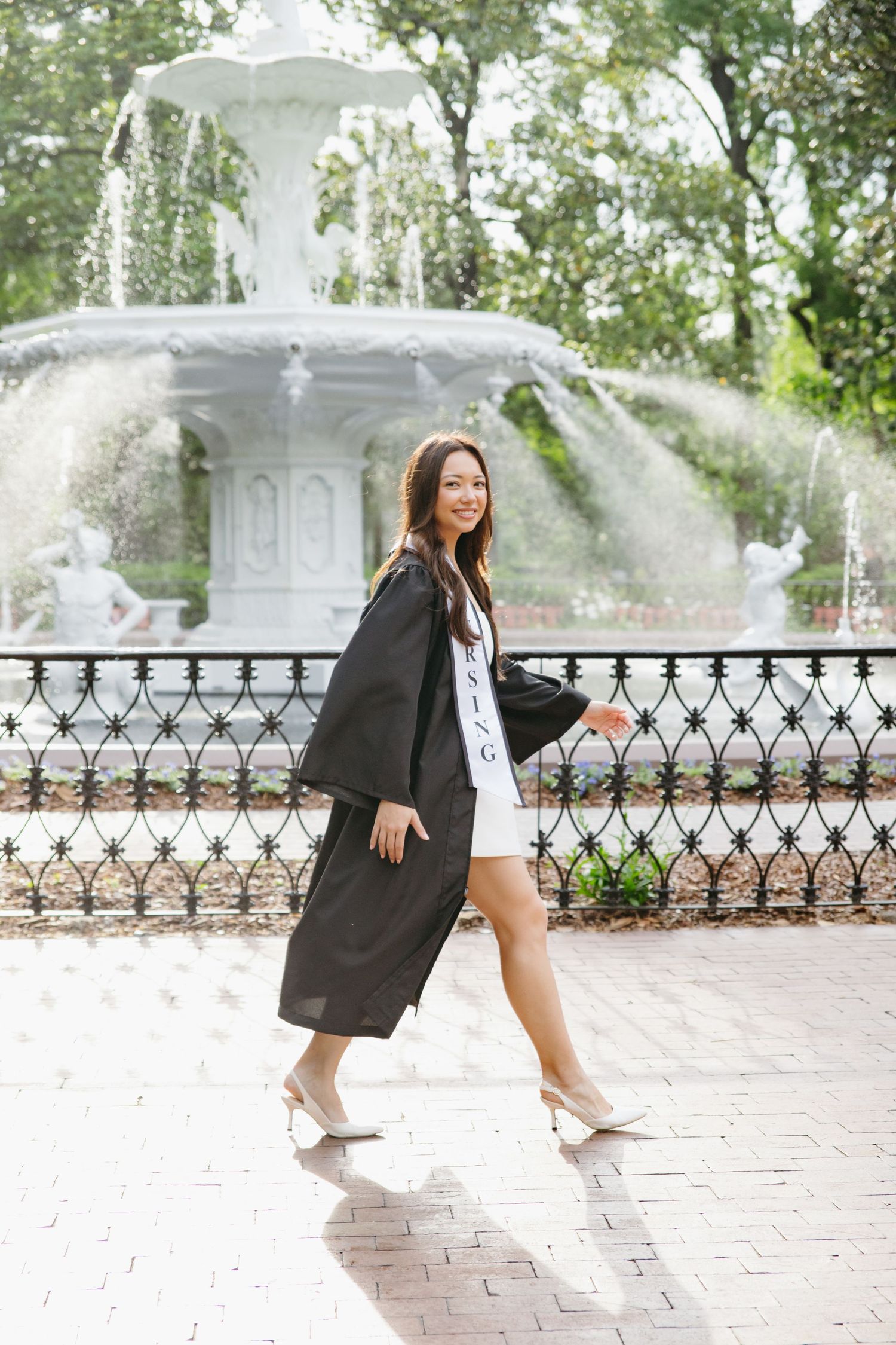 Nursing graduate in black gown walking near white fountain in scenic park setting.
