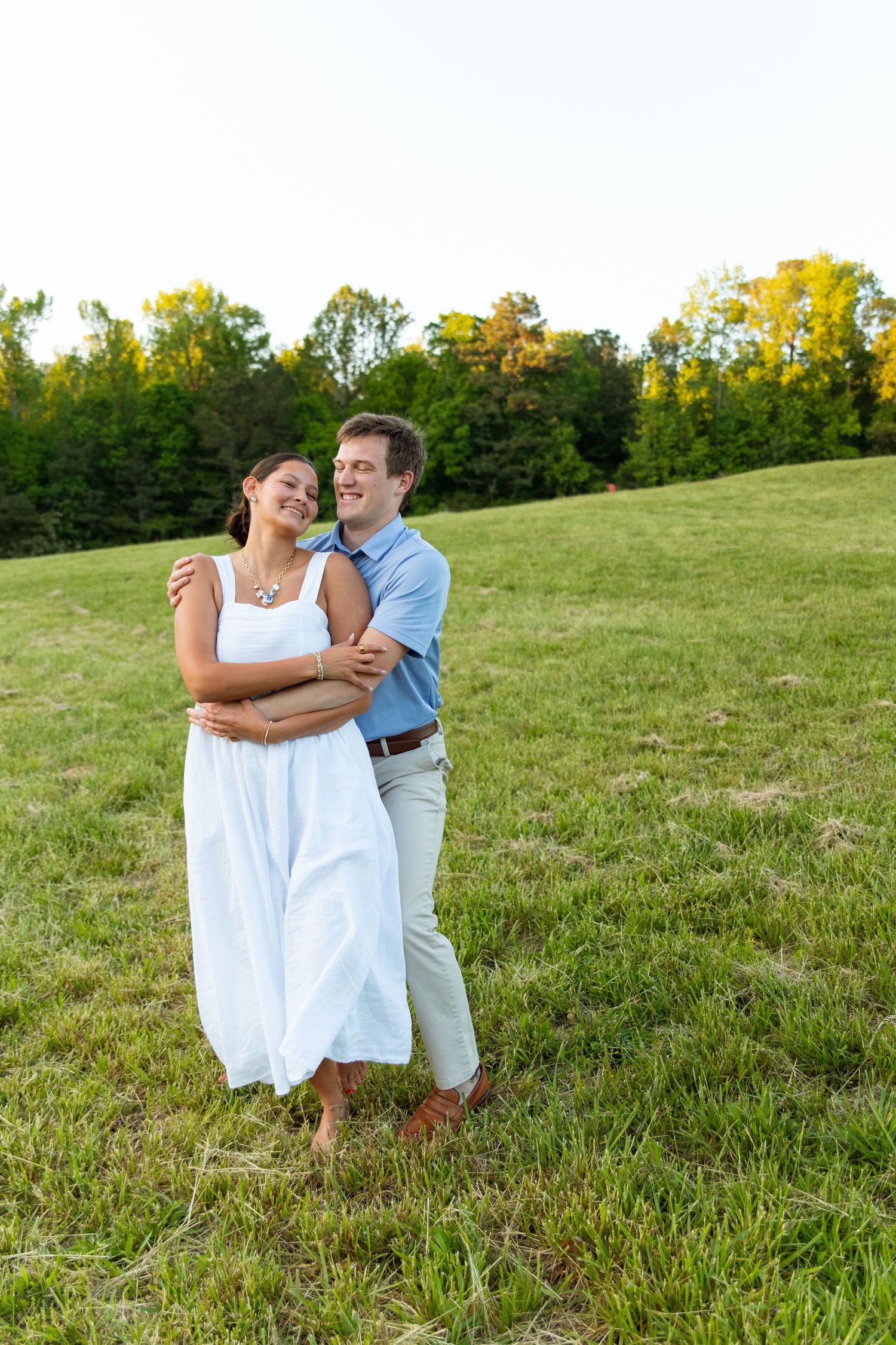 Happy couple hugging and laughing together on a green grassy hill at sunset with forest backdrop.