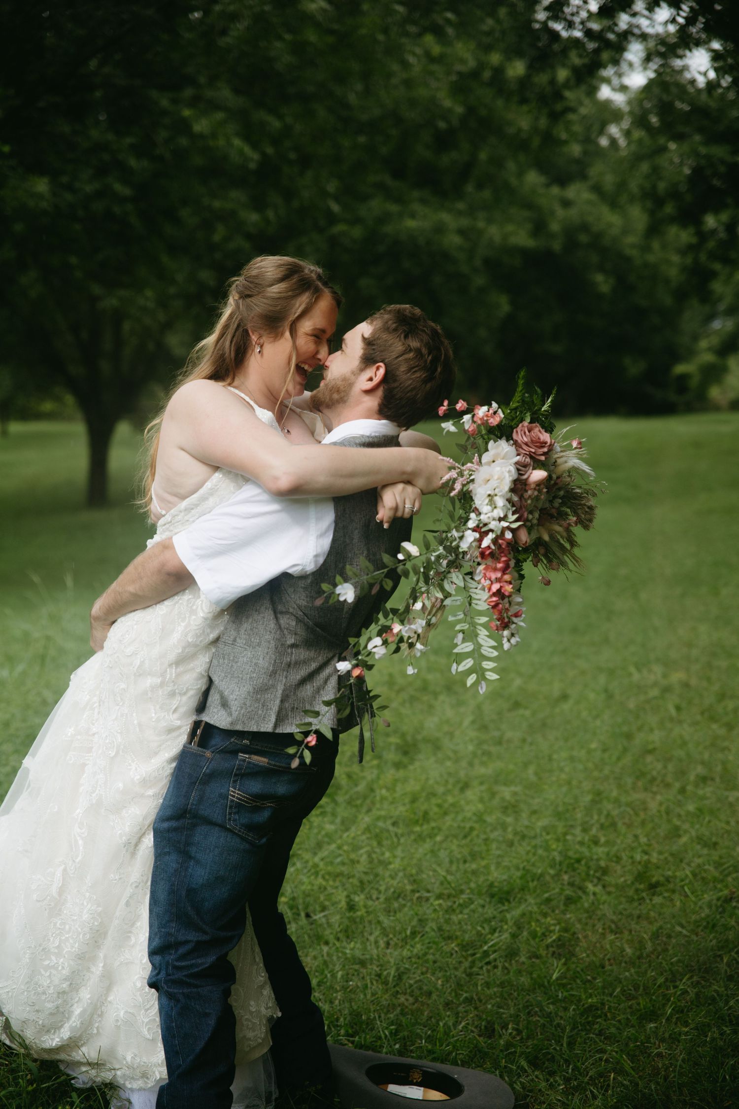 Groom lifting bride in a lace wedding dress while she holds a floral bouquet outdoors.