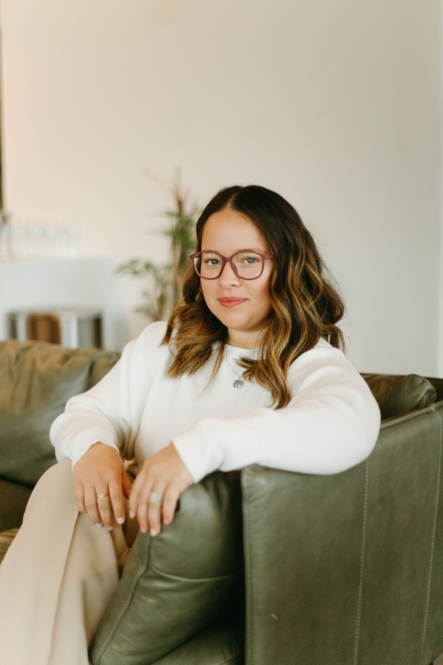 Young woman with glasses in white sweater sits relaxed on green leather sofa indoors.