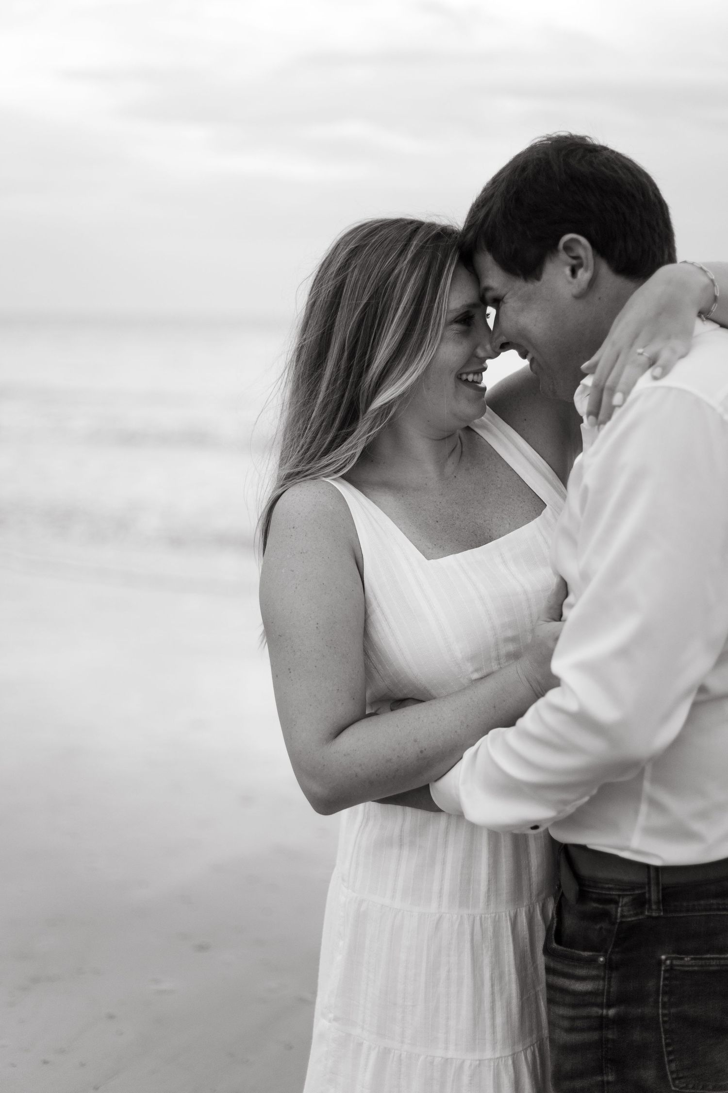 Black and white romantic couple embracing on a beach at sunset, smiling and touching foreheads together.