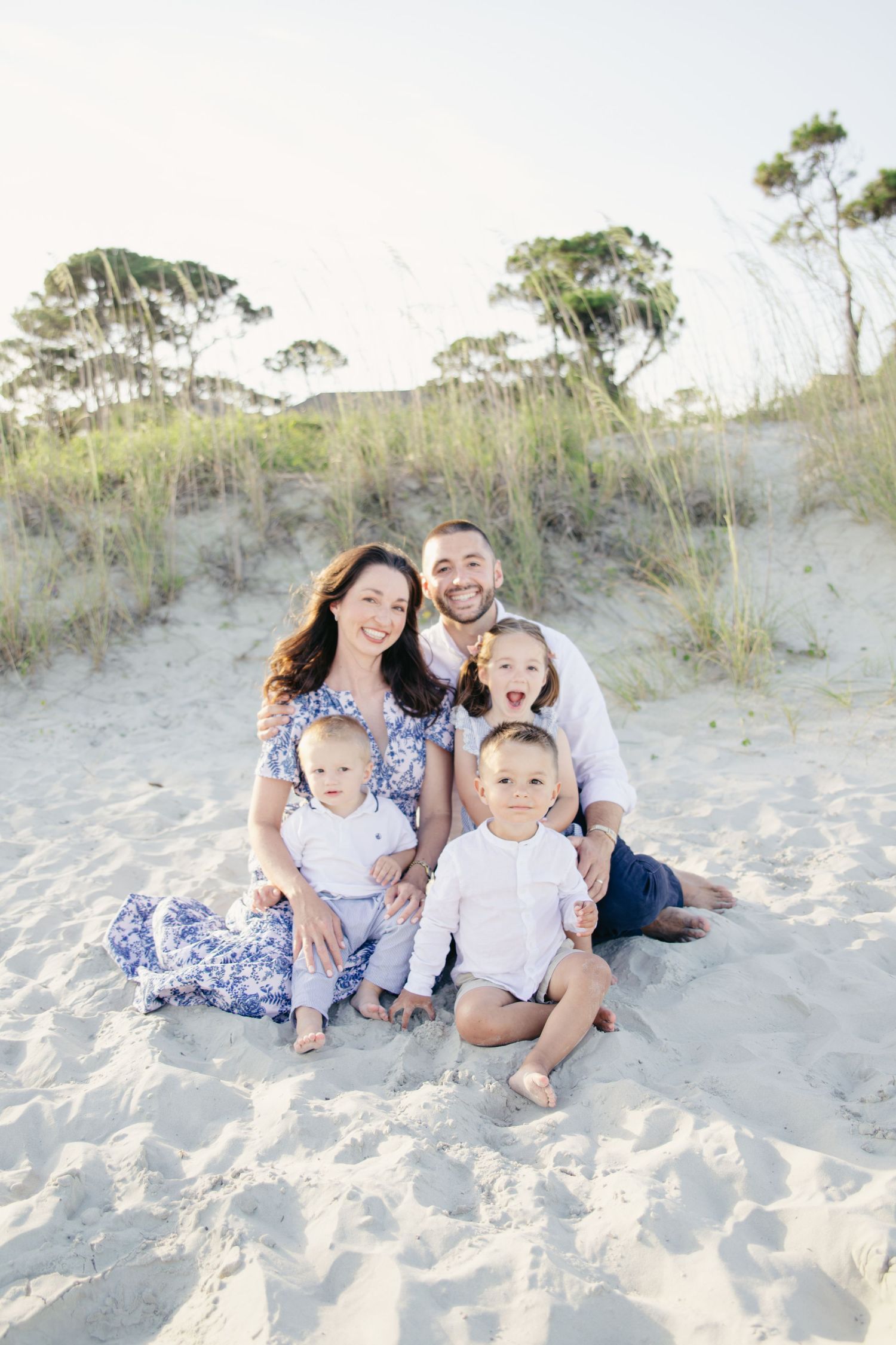 Happy family of five sitting on beach sand, parents with three young children smiling.